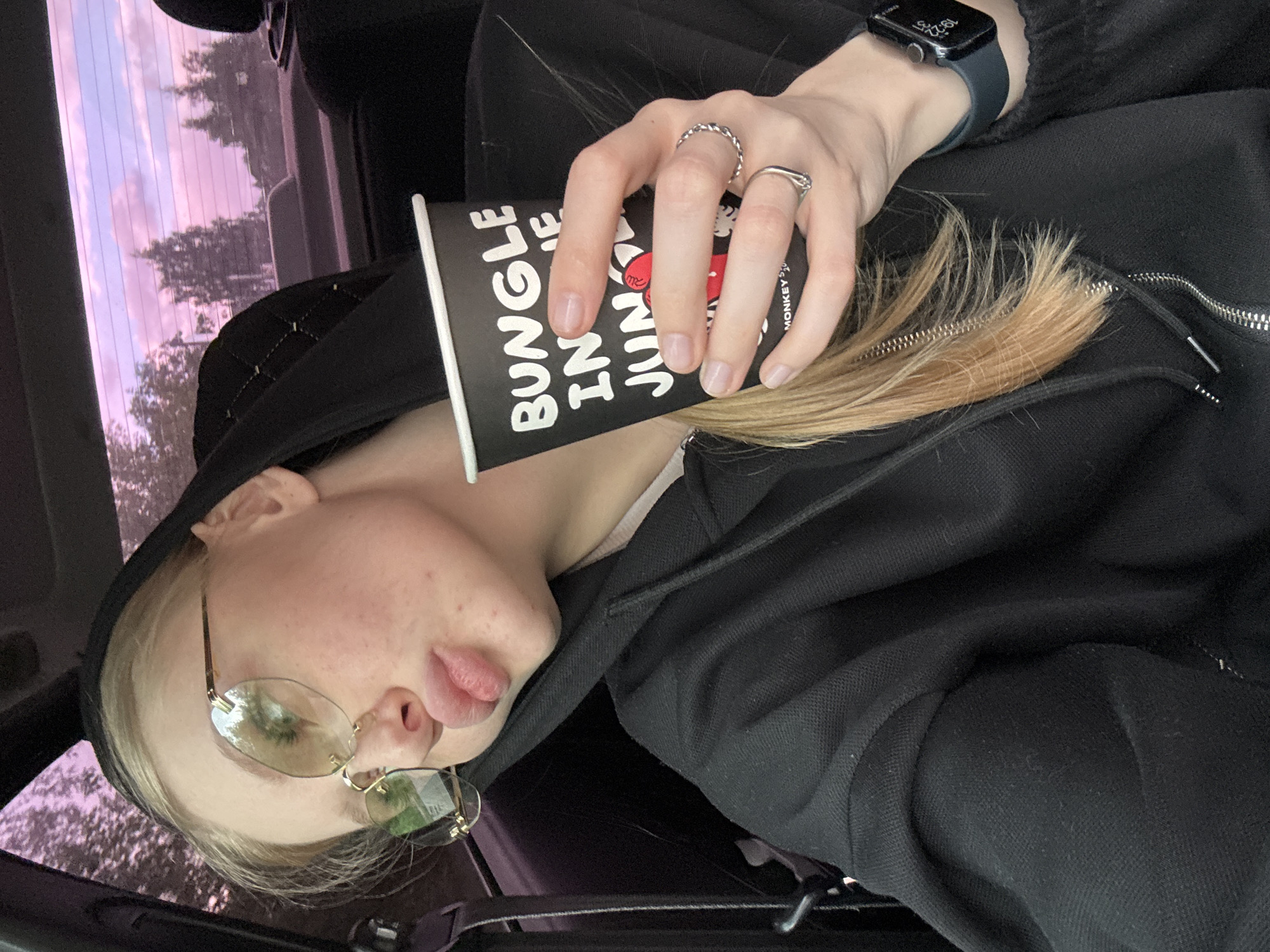 The image presents a selfie taken by a young woman inside a car. She is wearing a black hooded sweatshirt and gold-rimmed glasses, her blonde hair partially obscuring the zipper of her sweatshirt. She is holding a black paper cup with the words "BUNGLE IN JUNGLE MONKEY'S" printed on it, and is wearing a smart watch on her left wrist and two silver rings on her right hand. The background shows a blurred view of trees and a sky with pink and purple hues visible through the car window. The location is not determinable from this image.