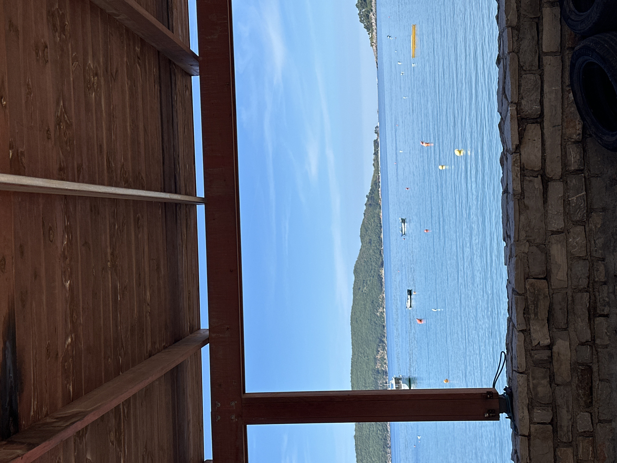 The image captures a view of the Adriatic Sea from what appears to be an enclosed structure. The scene is in Boreti, Montenegro. The perspective is partially obstructed by a wooden ceiling and support beams, suggesting the viewer is situated under a roof or within a building open to the water. Beyond the structure, the calm, blue waters of the Adriatic stretch towards the horizon, dotted with boats and buoys. The coastline features a lush green mountainous landscape that meets the sea. To the right of the frame, a stone wall and a stack of tires are visible. The overall impression is a tranquil coastal setting.