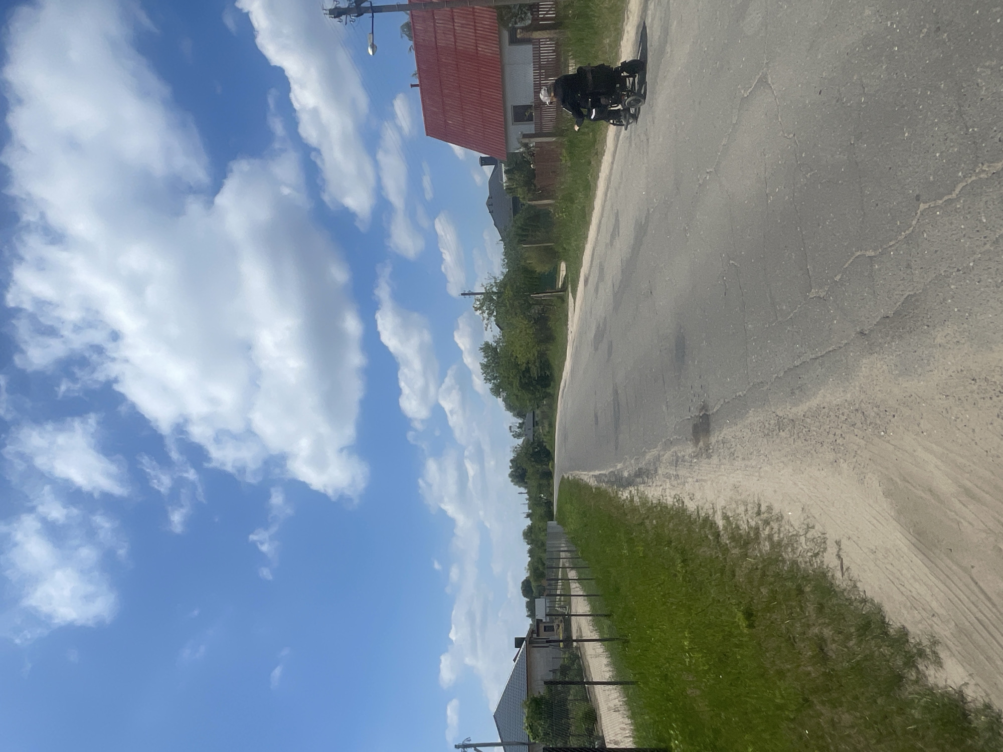 The media file shows a slightly tilted view of a rural road in Belarus, likely during the daytime on a sunny day with fluffy white clouds against a bright blue sky. On the right side of the image, a paved road stretches into the distance, bordered by a strip of sand and then lush green grass. A dark fence runs along the edge of the grass.
On the left side of the road, houses with red roofs and beige walls are visible, nestled amongst trees and vegetation. A utility pole with a light fixture stands prominently near one of the houses.
In the foreground, on the road, a person is riding a wheelchair, appearing to be moving along the road. The person is wearing dark clothing and a light-colored hat. The overall scene evokes a peaceful, everyday moment in a rural setting. There is no visible text in the image.