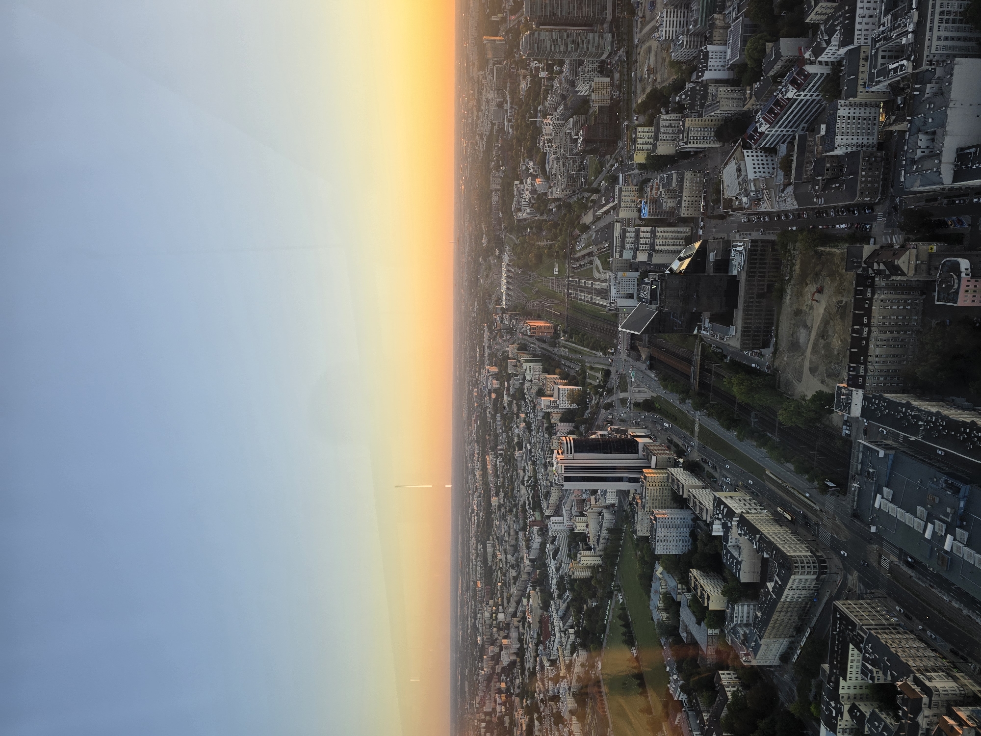 This is an aerial, eye-level, wide shot of a city landscape at dusk. The sky is a gradient of light blue at the top, transitioning to a soft orange glow near the horizon, suggesting the sun has recently set or is about to set. The city below is densely packed with buildings of varying heights, many appearing to be residential or office blocks. A major road runs through the center of the image, with cars visible on it, indicating it is a busy thoroughfare. There are also railway tracks visible, suggesting a significant transportation hub. In the foreground, there are several modern, tall buildings, some with reflective glass facades that catch the light of the sunset. In the background, the city stretches out to the horizon, with some structures faintly visible in the distance. The overall impression is of a sprawling urban environment bathed in the warm, fading light of day. The perspective suggests the photograph was taken from a high vantage point, possibly a skyscraper or tall building, within the city of Warsaw, Poland.