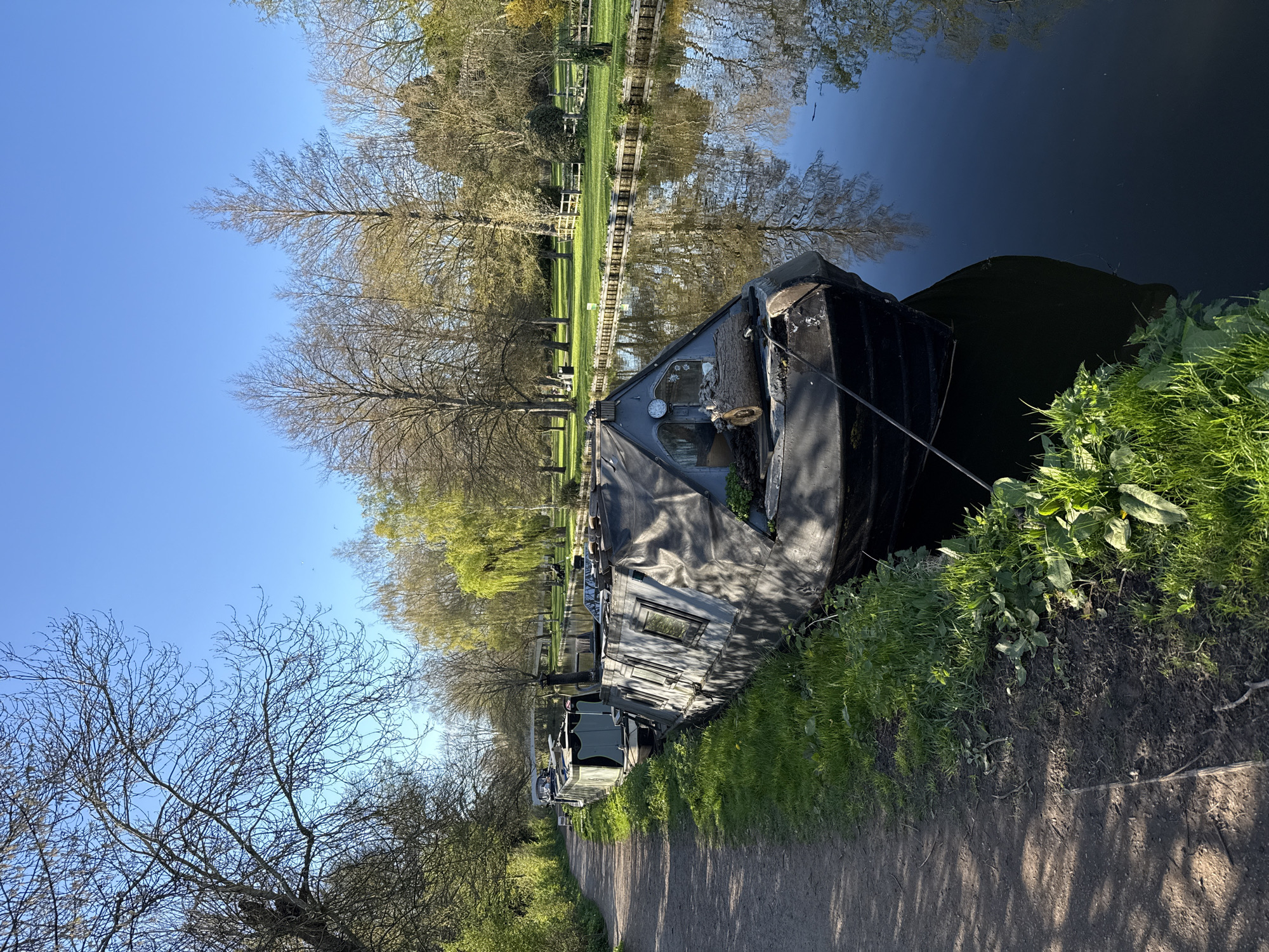 A horizontal, eye-level shot shows a canal scene likely within Dacorum, United Kingdom. A long, narrow black canal boat is moored alongside a towpath. The towpath is a dirt path, and vegetation grows between the boat and the path. The canal water is dark and reflects the trees and sky on the opposite bank. Trees line the canal, some bare, others with emerging green leaves. The sky is a clear, bright blue.