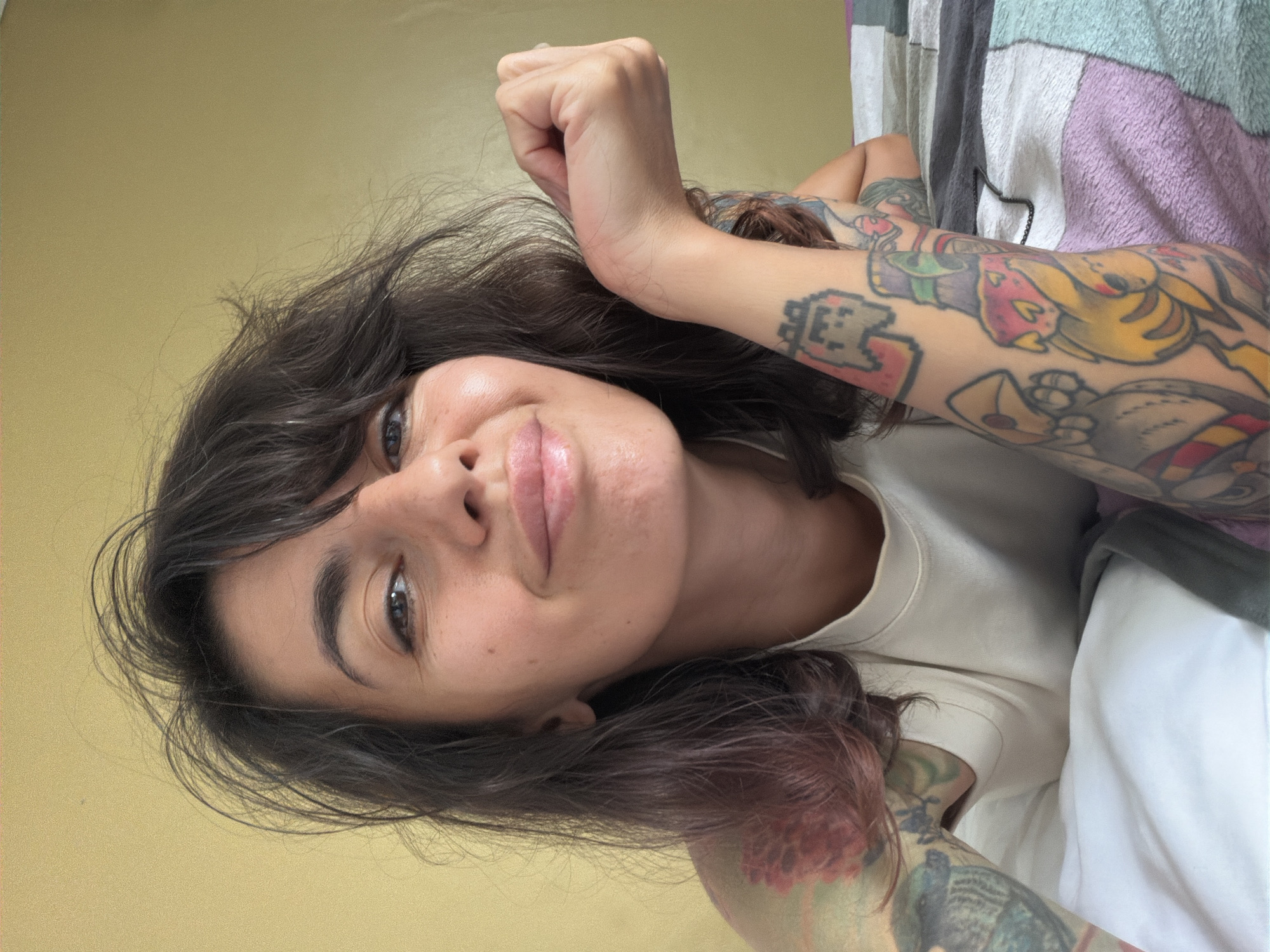 The image depicts a young adult female reclining indoors in Kuta Selatan, Indonesia. She is positioned facing the viewer, with her upper torso and head visible. Her dark brown hair is styled with bangs, appearing slightly tousled. Her eyes are dark, and she exhibits a subtle smile with full, glossy lips. The woman wears a light-colored, possibly off-white, short-sleeved top with a rounded neckline. Both of her forearms and upper arms are extensively covered with colorful, cartoon-style tattoos; notable designs include a grey pixelated ghost and a yellow character with red accents on her right arm (viewer's left), and a large floral design on her left shoulder (viewer's right). Her left hand is resting on her right forearm. She is lying on a bed, partially covered by a white sheet or pillow on the left side, and a multi-colored blanket or bedspread on the right. The blanket features sections of white, dark grey, light grey, and lavender. A thin, dark chain is visible on the blanket near her right elbow. The background consists of a plain, light yellow wall. The scene is illuminated by ambient light, creating soft highlights on her face.