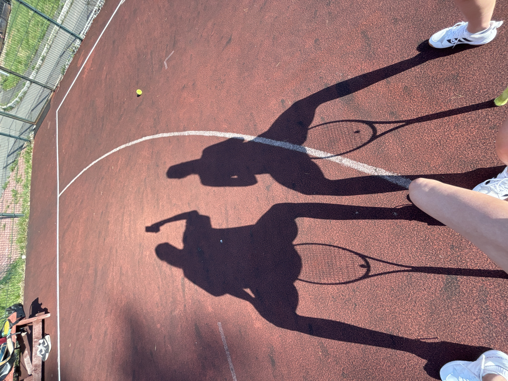 The image shows a tennis court with a red surface and white lines. Shadows of two people holding tennis rackets are visible on the court. A tennis ball is near the line on the left side of the image. The edge of the court is visible on the left with green grass and a fence. In the lower left corner, a bench with tennis equipment and shoes is present. Partial views of the players' shoes and legs appear on the right. The location is likely Прикамский, Russia, based on provided information, but this cannot be confirmed solely from the image.