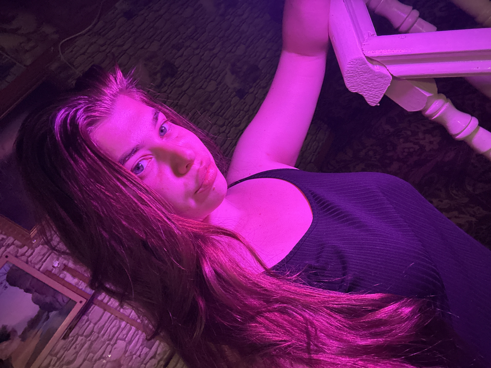 The image shows a young woman with long, dark hair illuminated by pink light. She is wearing a black, ribbed tank top and is leaning on a white wooden railing. The background consists of a stone wall with a framed picture and a patterned carpet. The overall setting appears to be indoors, possibly a staircase or hallway, with artificial lighting creating a vibrant pink glow. No specific location in Мещёры, Russia, can be identified from the image.