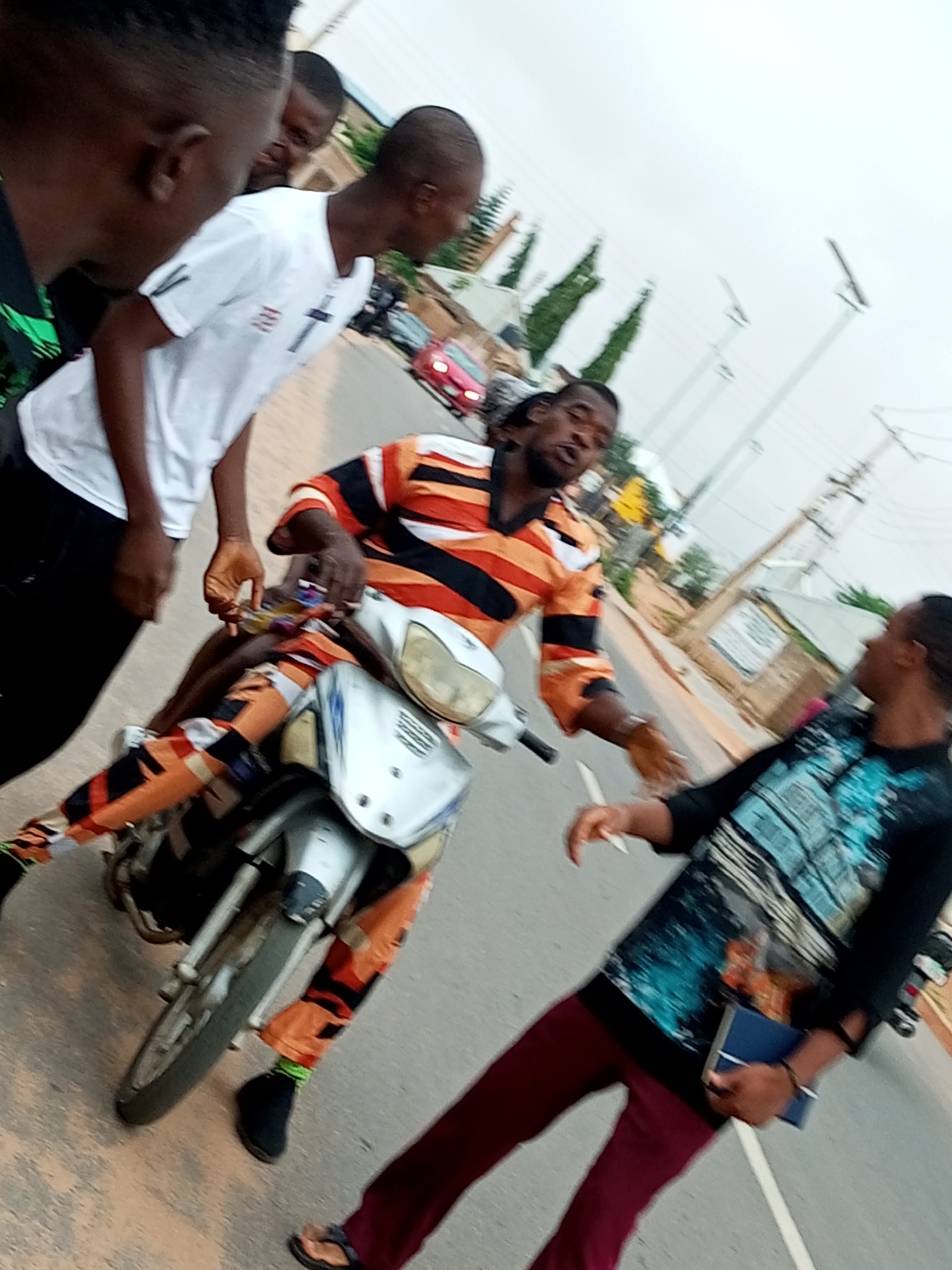 In Gombe, Nigeria, a group of young men are gathered around a motorcycle on an asphalt road. The central figure, seated on the motorcycle, is wearing a brightly colored striped shirt and matching pants, with a gold chain around his neck. He is speaking or shouting with his mouth open, gesturing with his hands. To his left, another young man in a white t-shirt with a cross emblem is leaning forward, looking towards the seated individual. Behind them, other men are partially visible. The environment suggests a street in Gombe, with buildings, trees, and utility poles visible in the background. The overall atmosphere appears to be energetic, possibly a social gathering or a street scene.