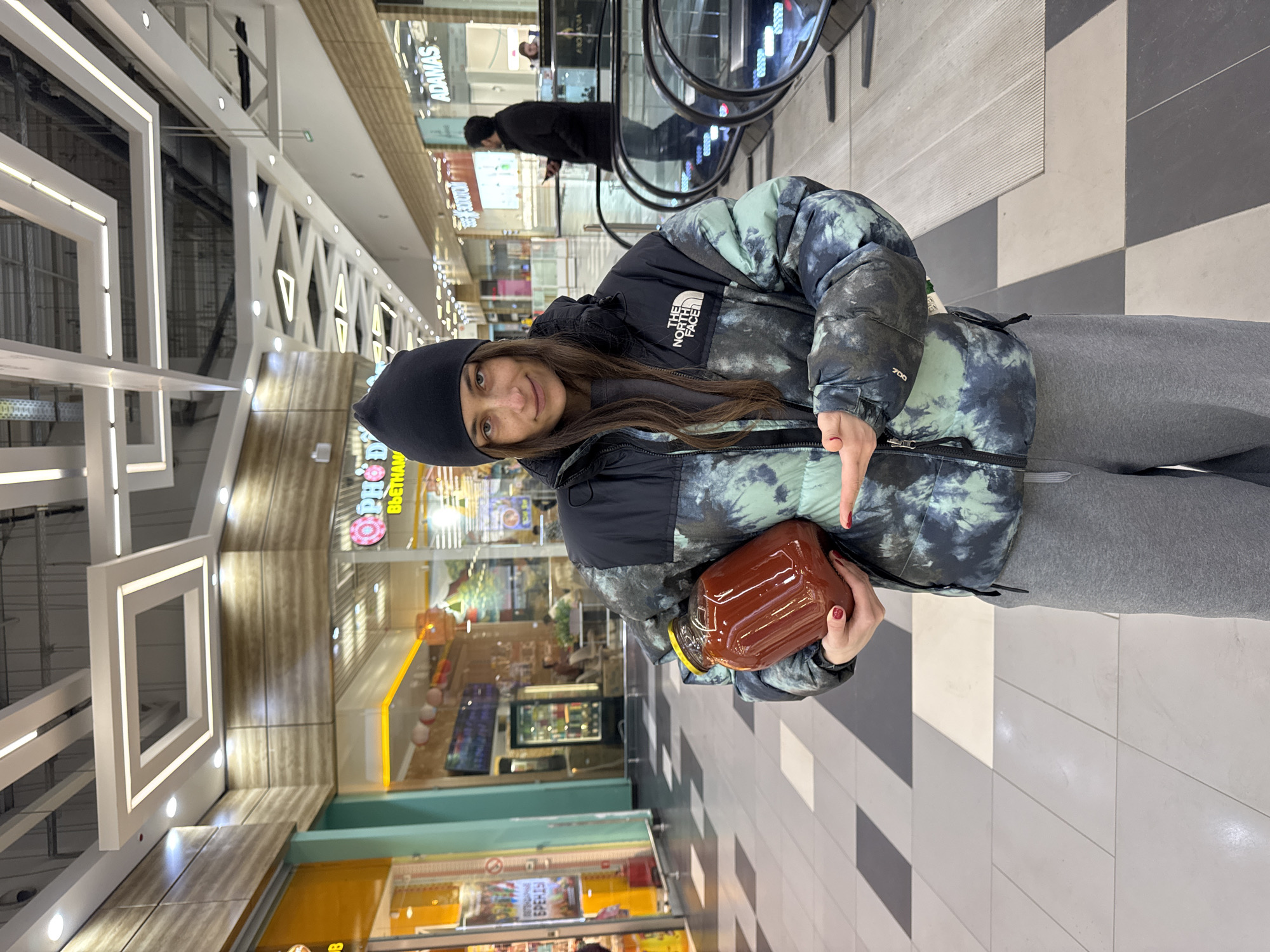 A woman is indoors, likely in a shopping mall in Moscow, Russia. She wears a black beanie, a patterned puffer jacket with "THE NORTH FACE" visible, and gray sweatpants. She holds a large, dark-colored jar with a gold lid in her hands. The background shows retail stores, an escalator, and modern lighting fixtures. The floor has a striped pattern of light and dark tiles. The woman points to the jar with her right hand.