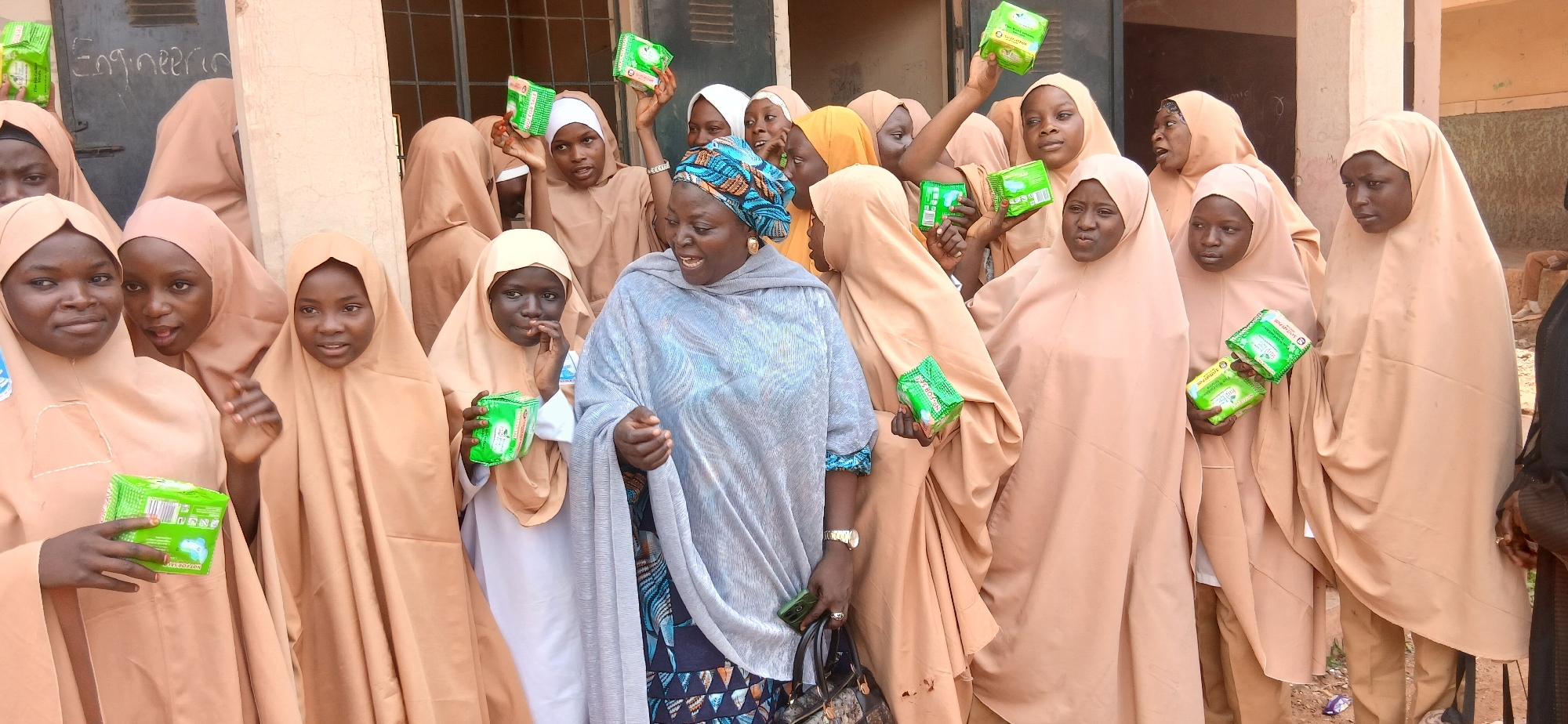 A group of young girls and a woman are gathered outdoors, possibly in a schoolyard in Unguwar Shanu, Nigeria. The girls are all wearing light brown hijabs and dresses, while the woman is wearing a light blue hijab and a patterned dress. They are holding up small green packages, which appear to be sanitary pads. Some of the girls are smiling and looking towards the camera, while others seem to be engaged in conversation. The overall mood appears to be positive and celebratory.
In the background, there is a building with a window and a door. There is also some writing on the wall next to the door that says "Engineering". The ground is covered in dirt and some debris. The lighting suggests it is daytime, likely sunny. The focus is on the people and the sanitary pads they are holding. The event seems to be related to a distribution or awareness campaign about menstrual hygiene.