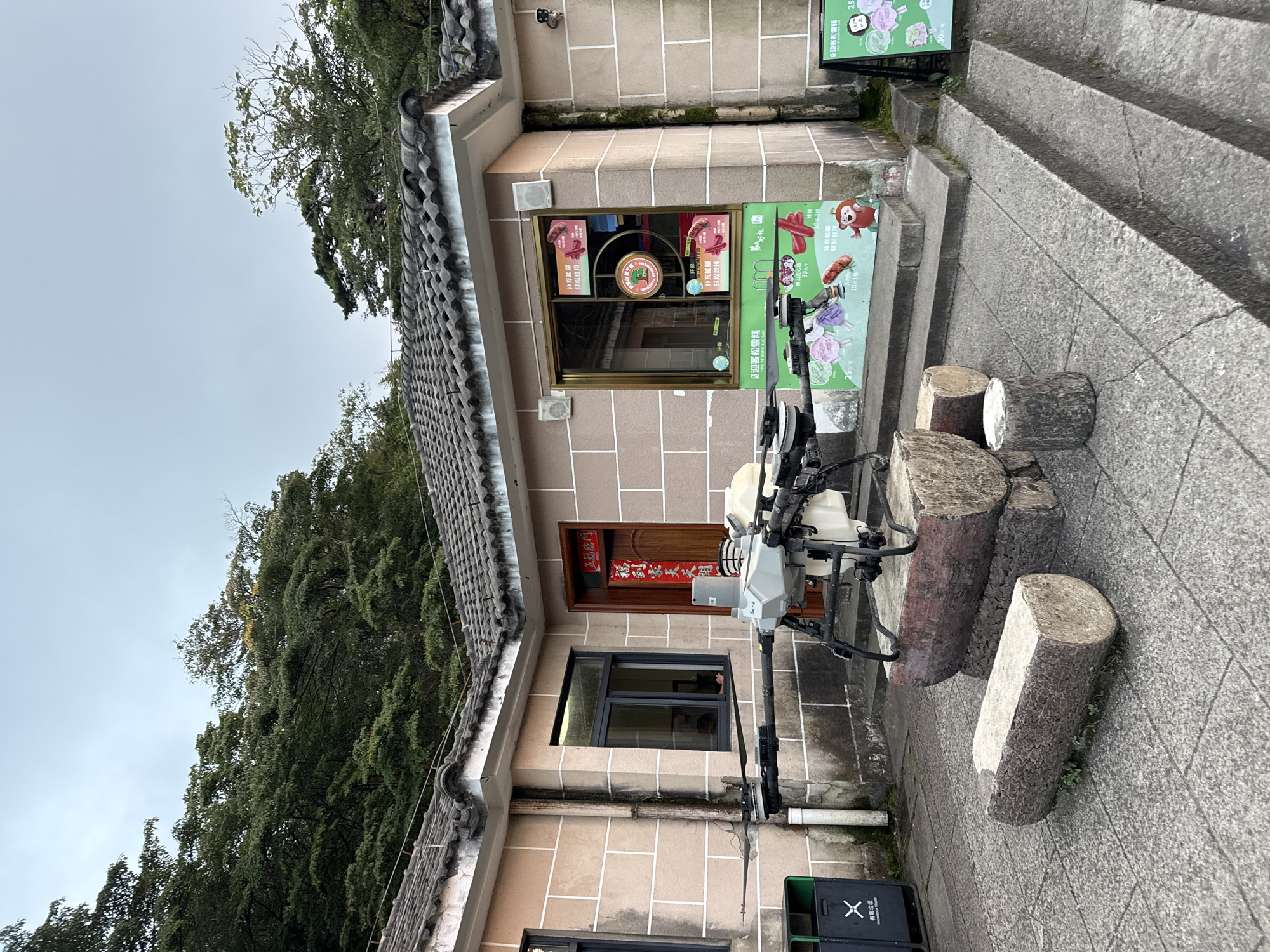 The image depicts an outdoor scene in Huangshan District, China. In the foreground, a large drone is positioned on a base made of several logs. The drone has multiple propellers and a white tank. To the left of the drone, a dark green trash bin with a white "X" logo is visible. Behind the drone and logs, a light-colored, stone-faced building with a tiled roof is situated. The building has a window and a wooden door with red Chinese calligraphy on it. To the right of the building, a glass-paned service window displays several advertisements, including images of food items and cartoon characters. A set of concrete steps leads up to the service window, with a green sign on an easel to the right of the steps. The background shows a dense area of green trees and vegetation, with a cloudy sky above. The overall environment suggests a somewhat rural or scenic location, possibly a rest stop or service area.