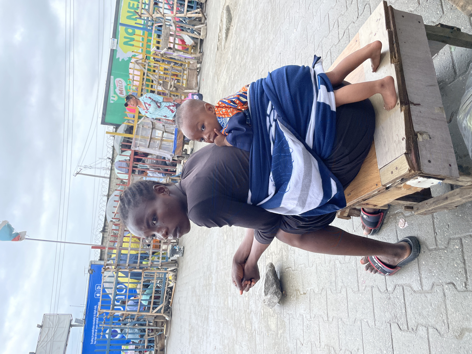 A young Black woman wearing a dark brown, long-sleeved dress sits on a wooden bench with her back to the camera in an outdoor market in Lekki, Nigeria. She is holding her hands clasped in front of her on her lap. A young Black child is strapped to her back with a blue and white striped cloth. The child is wearing a colorful, patterned shirt and has a finger in their mouth. The ground is paved with gray rectangular stones, and it appears to be wet. To the left of the bench, a gray rock rests on the pavement. In the background, there are various stalls and structures made of wood and metal, some with colorful signs and fences. A flag with red, white, and blue colors flies in the upper left corner of the frame. The sky is overcast and gray. Several people can be seen in the background, some standing and others behind structures. A prominent green billboard with "MILO" written on it is visible in the mid-ground.