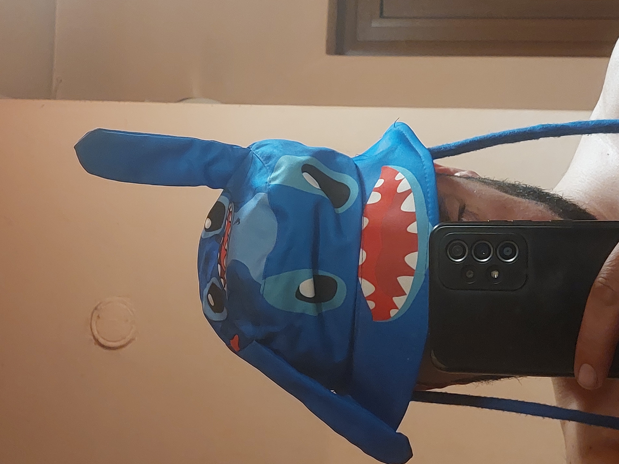 The image depicts a person holding a black smartphone in front of them, partially obscuring their face, and wearing a blue plush hat shaped like the Disney character Stitch. The hat has prominent ears, a mouth with red inside and white teeth, and facial features resembling the character. The person appears to be taking a selfie or a photo in a room with light beige walls. There is a round object, possibly a light fixture, on the wall. A window frame is visible in the background. No information is available to suggest that the image was taken in Tsarevo, Bulgaria, and this cannot be confirmed.