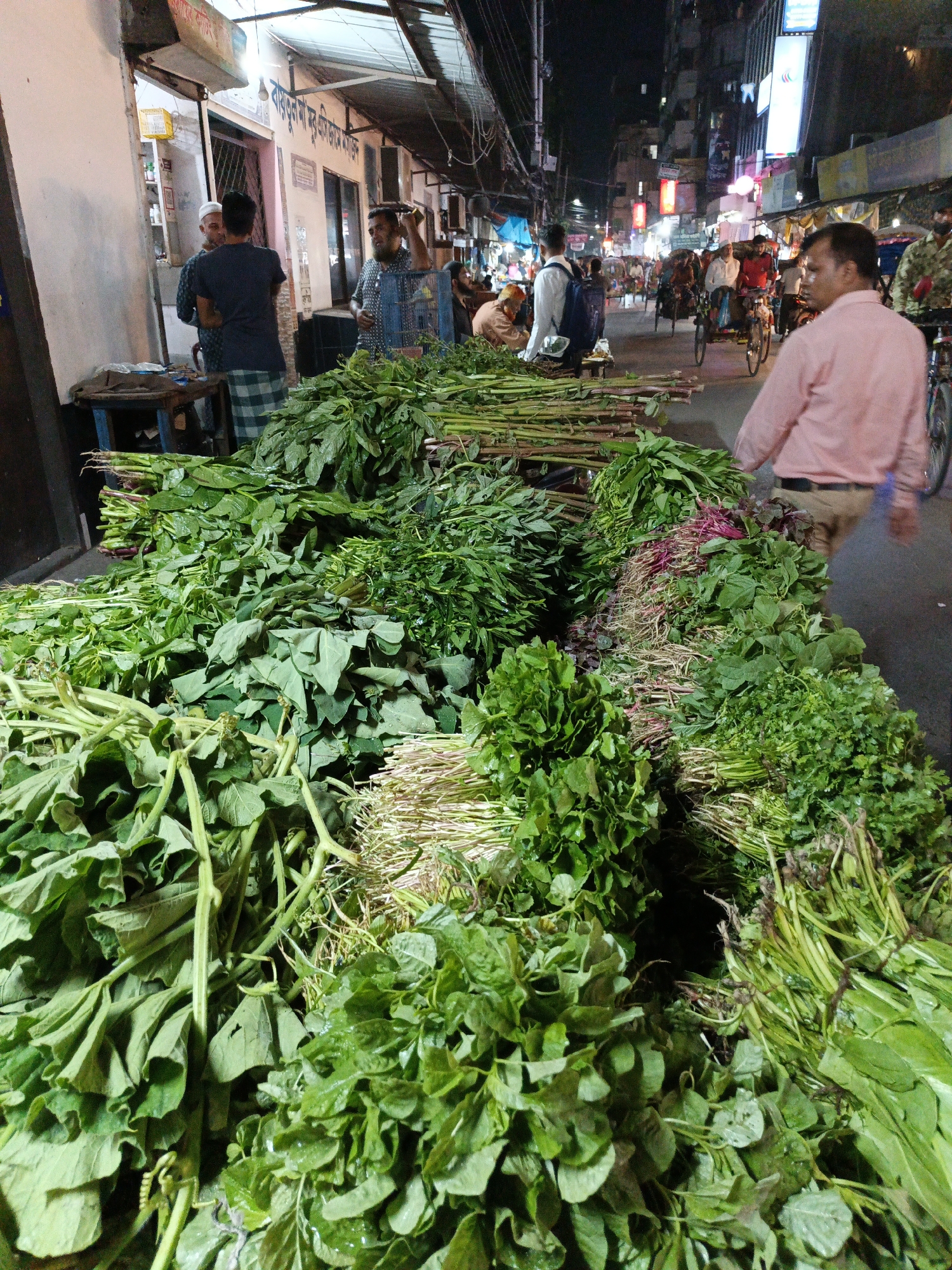 The image depicts a bustling street scene in Dhaka, Bangladesh. Heaps of various green leafy vegetables are prominently displayed, seemingly for sale on the roadside. Numerous people populate the scene, including potential customers and vendors. There's a person wearing a pink shirt walking past the vegetables. In the background, rickshaws and cyclists navigate the crowded street, illuminated by a mix of streetlights and shop lights. The overall atmosphere suggests a vibrant and active night market. Shop signs are visible in Bengali script.