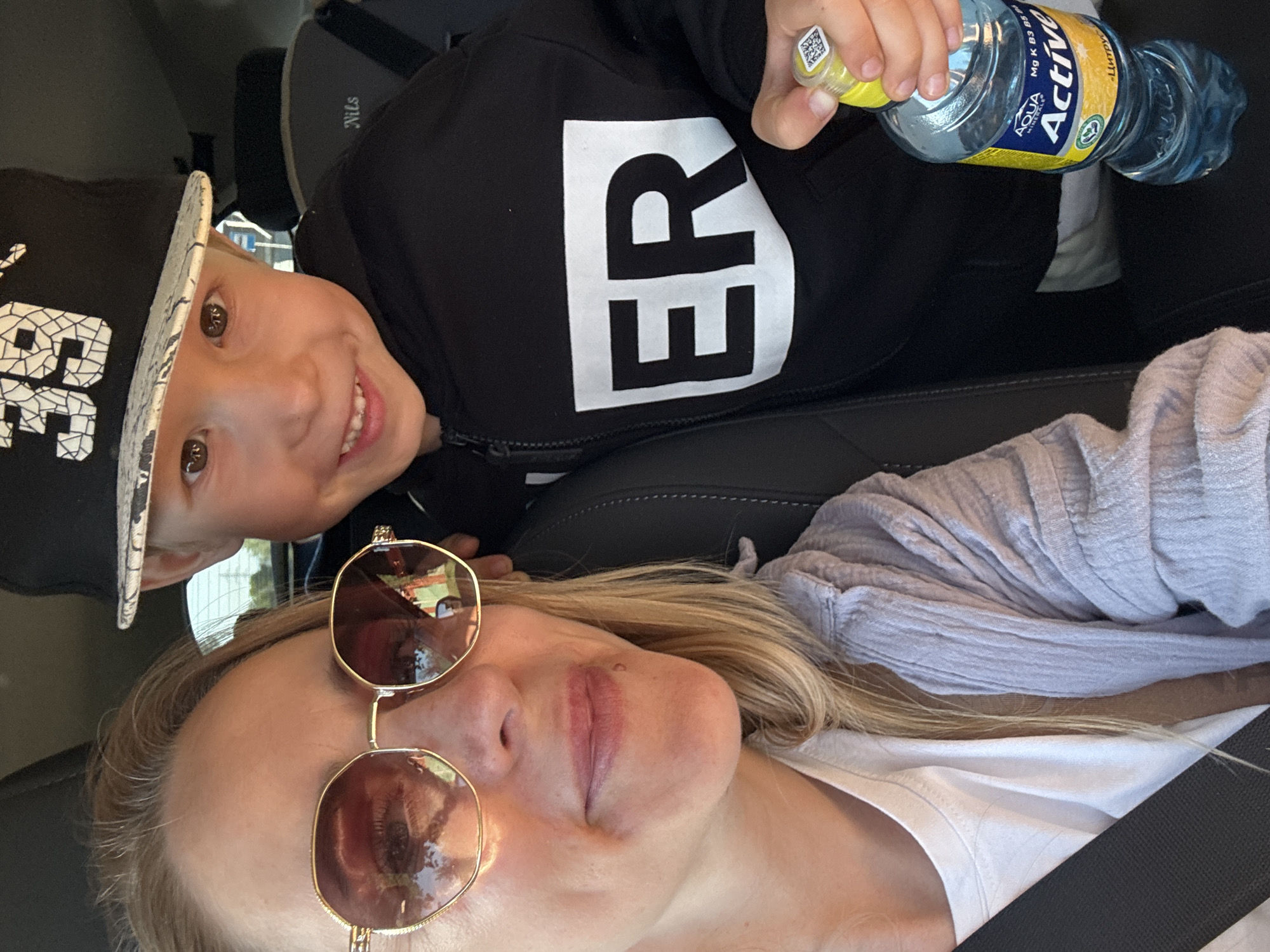 A selfie taken indoors, likely in a car. A woman with blonde hair, wearing sunglasses, is seated with a seatbelt visible across her chest. A child wearing a black hoodie with white lettering is leaning over her. The child is holding a bottle of "Aqua Active" water. The location is not clearly discernable as Chaikovsky, Russia from the image.
