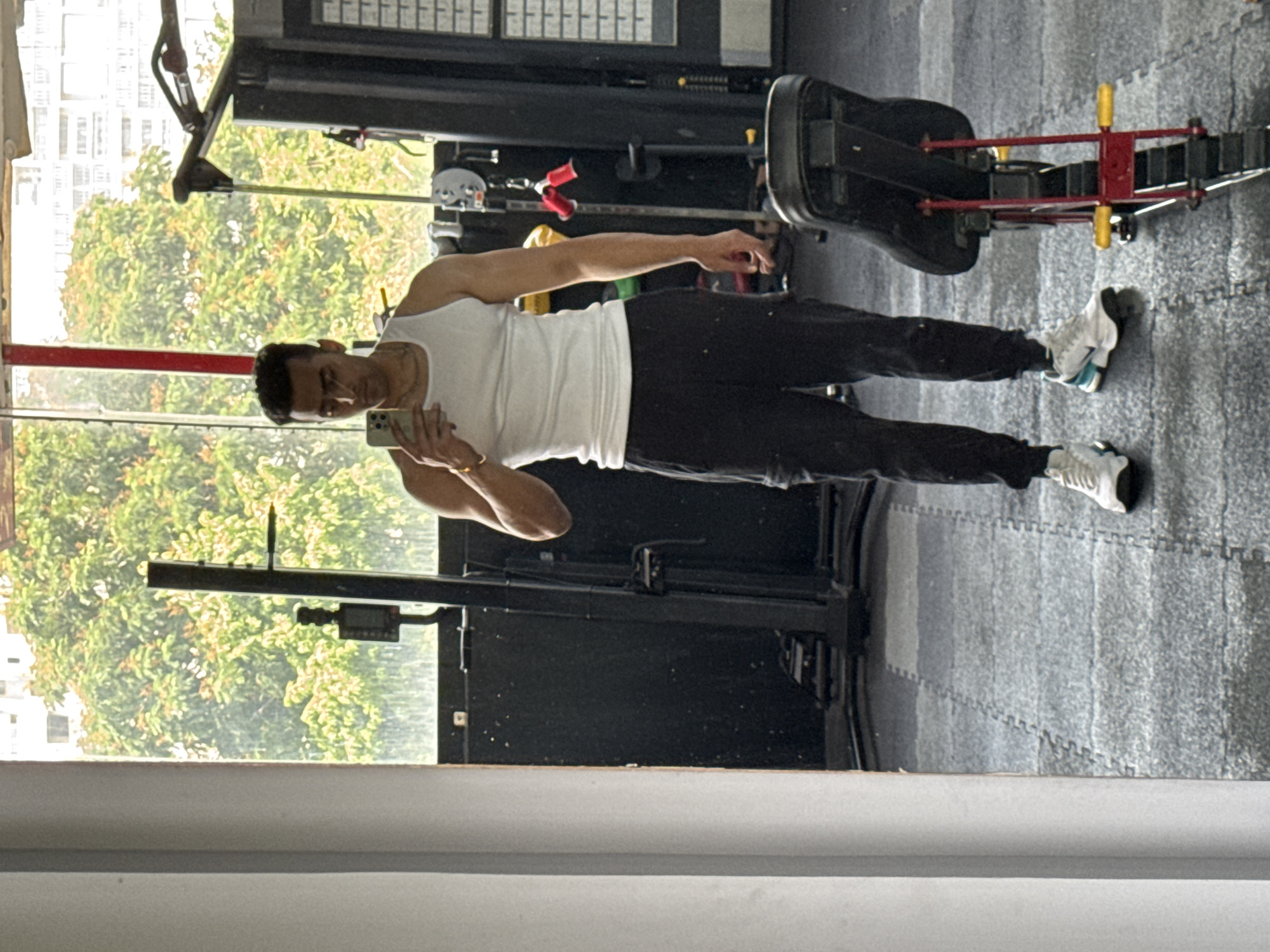 A man is taking a mirror selfie in a gym. The man is wearing a white tank top and black pants and white sneakers. He is holding a cell phone in his left hand. The gym has workout equipment. The location is suggested as Kolkata, India. Through a window in the gym, there are trees and buildings visible.