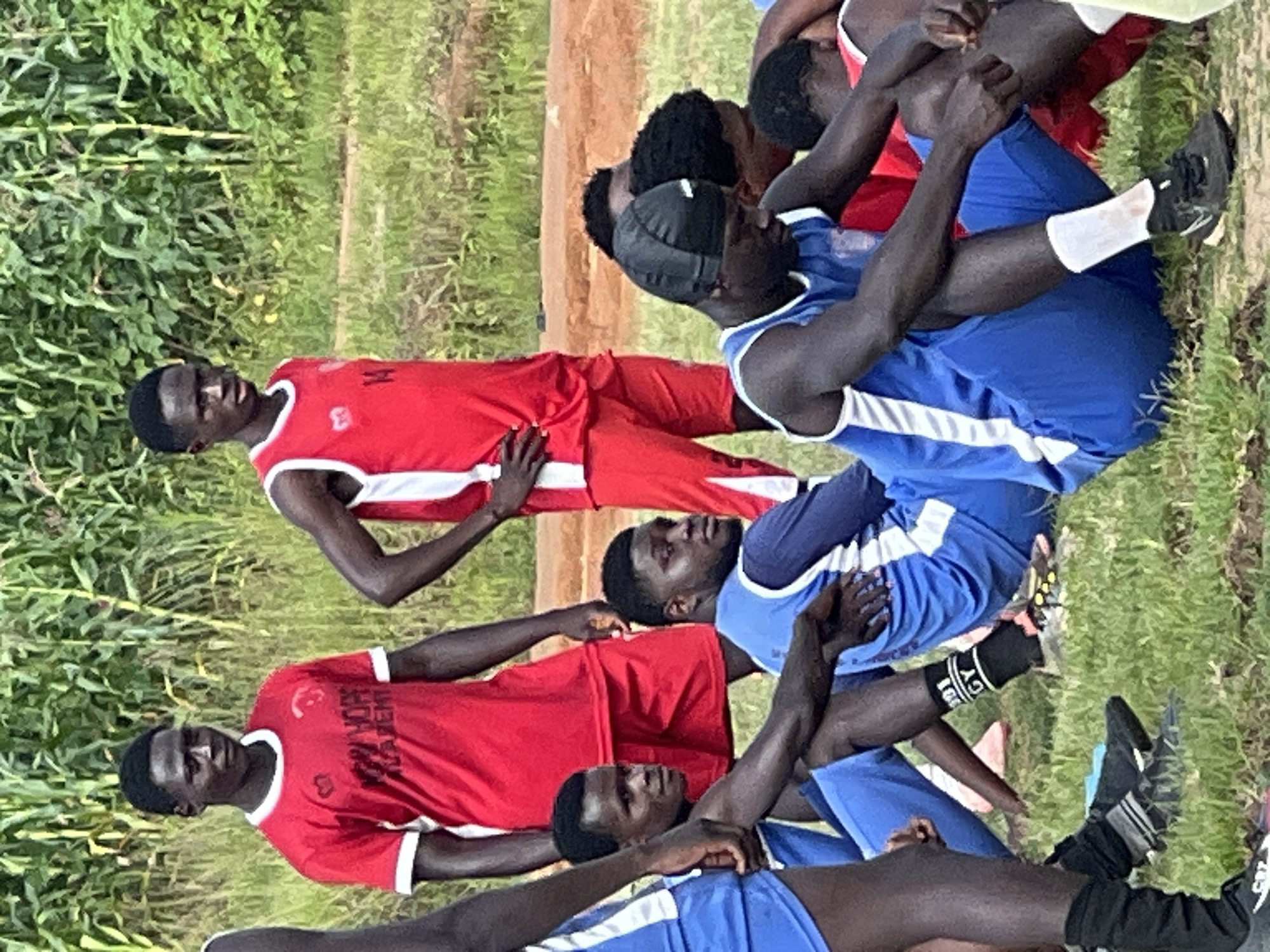 This image depicts a group of young men, presumably athletes, gathered outdoors in what appears to be a grassy field. The background is dominated by tall green plants, possibly corn stalks, suggesting a rural or semi-rural environment. The ground is covered in a mix of dry grass and reddish-brown soil.
Several individuals are visible, some standing and others sitting or kneeling. They are wearing athletic attire. Two individuals in the background are wearing red tops with white trim, and one has "NEW HOPE ACADEMY" printed on his shirt. In the foreground and middle ground, several individuals are wearing blue athletic tops and shorts. One person is wearing a black skullcap. Some are looking towards the camera, while others are looking away or downwards. The overall impression is of a sports team or group taking a break during training or a match.
While the image itself does not explicitly contain elements that confirm its location in Jos, Nigeria, the context of athletic activities and the depicted environment are consistent with such a location.