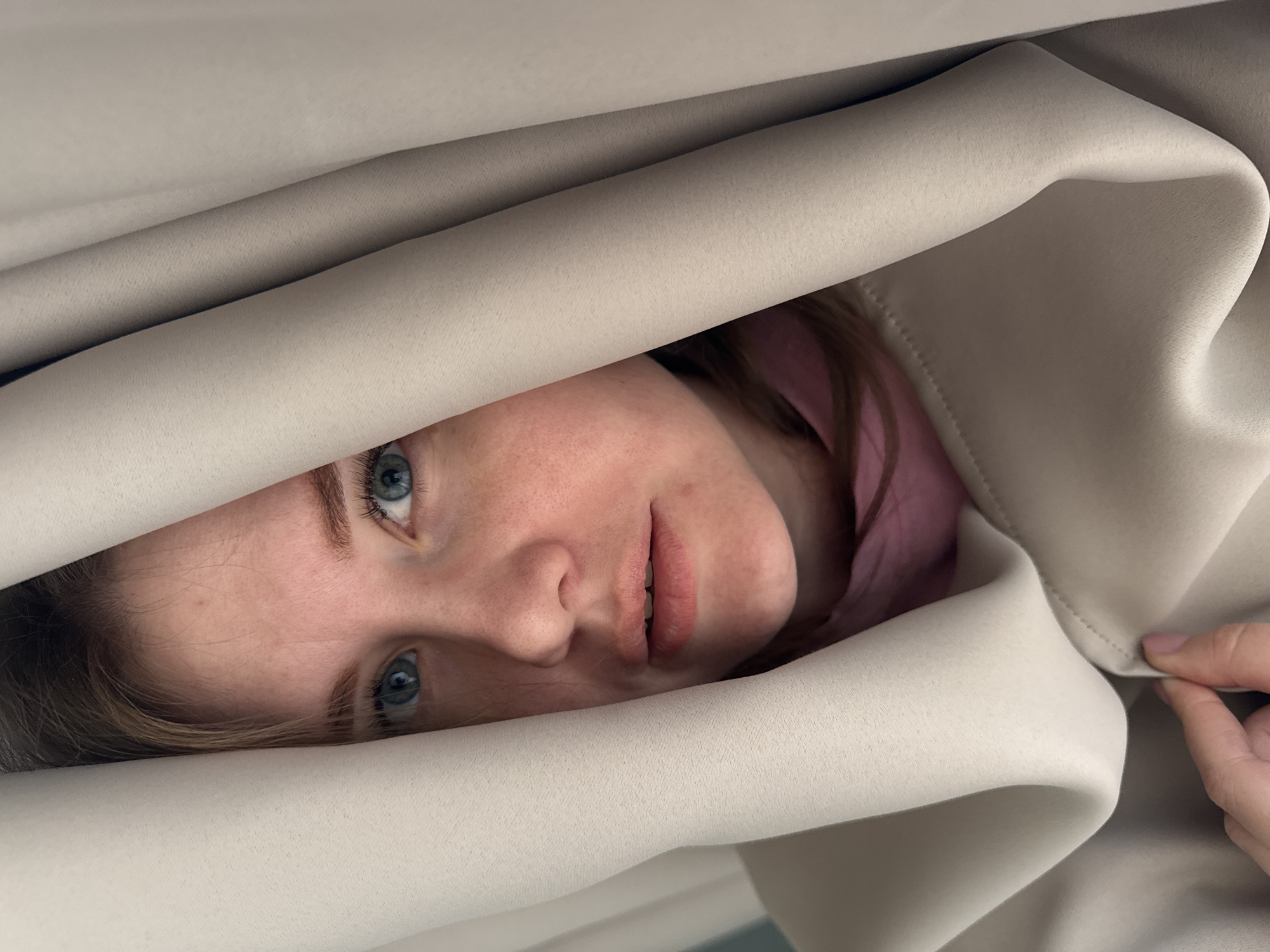 This vertical close-up image, captured in Gornyi (Dvurechenskoe S/P), Russia, depicts the upper portion of a female subject, partially obscured by light beige fabric. The individual's face, featuring light skin, blue eyes, and light brown hair visible at the top, is framed centrally by two vertical folds of the beige material. Her gaze is directed straight forward, and her mouth is slightly open. A pink garment is visible around her neck and chest area, beneath the beige outer layer. A hand, exhibiting visible fingernails, is positioned in the lower left quadrant of the frame, gripping the beige fabric which displays a stitched seam. The immediate background is entirely obscured by the subject and the surrounding fabric, with a minor, indistinct light blue element visible in the upper left corner.