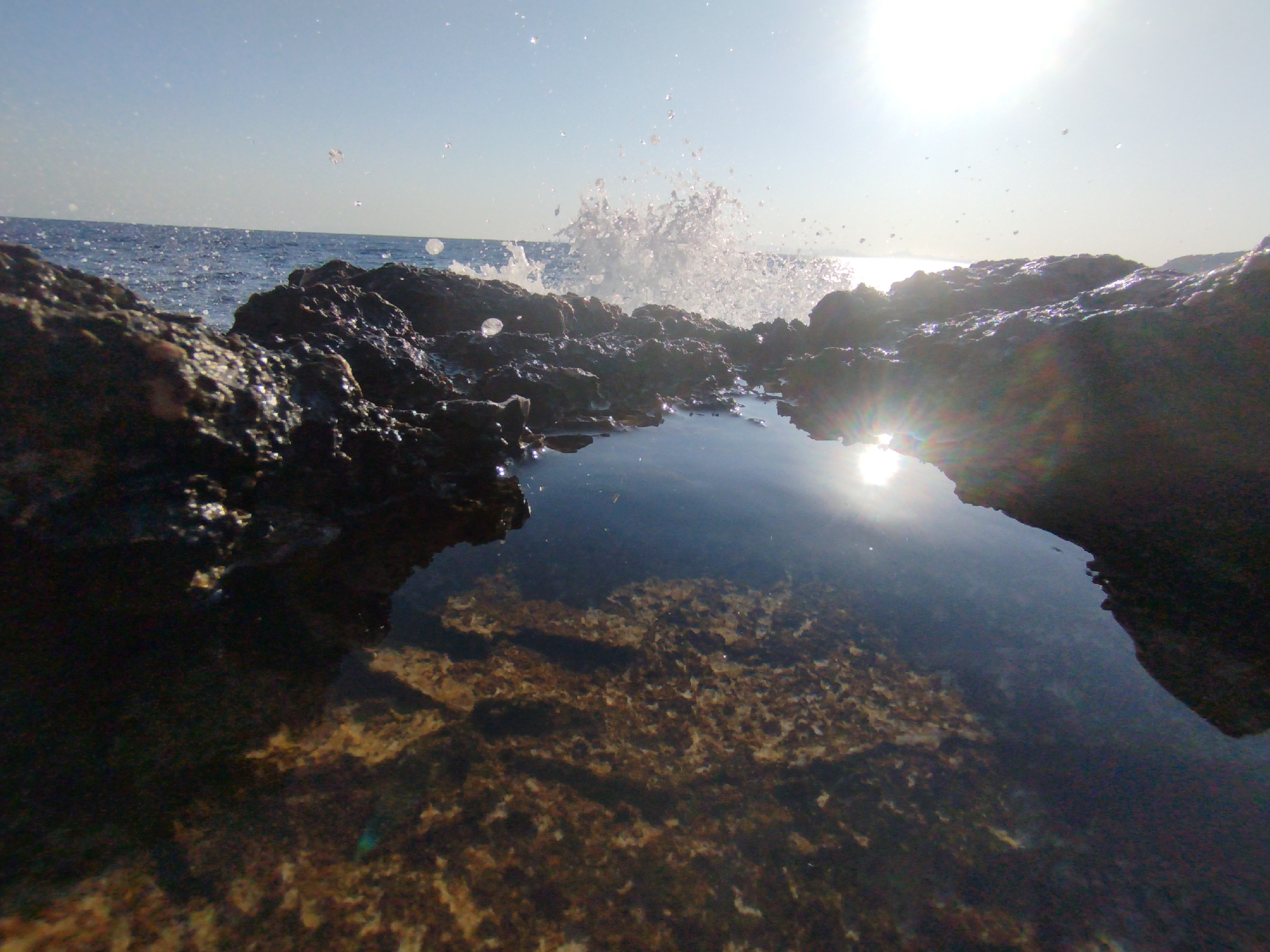 A low-angle, wide-shot captures a natural rock tide pool with clear water, revealing a textured bottom, in Karaburun, Turkey. The sun, a bright lens flare in the upper right quadrant, casts a strong reflection on the water's surface. Jagged, dark rocks frame the foreground and middle ground. In the background, a wave crashes against the rocks, sending a spray of water into the air. The ocean stretches to the horizon under a clear, bright sky.