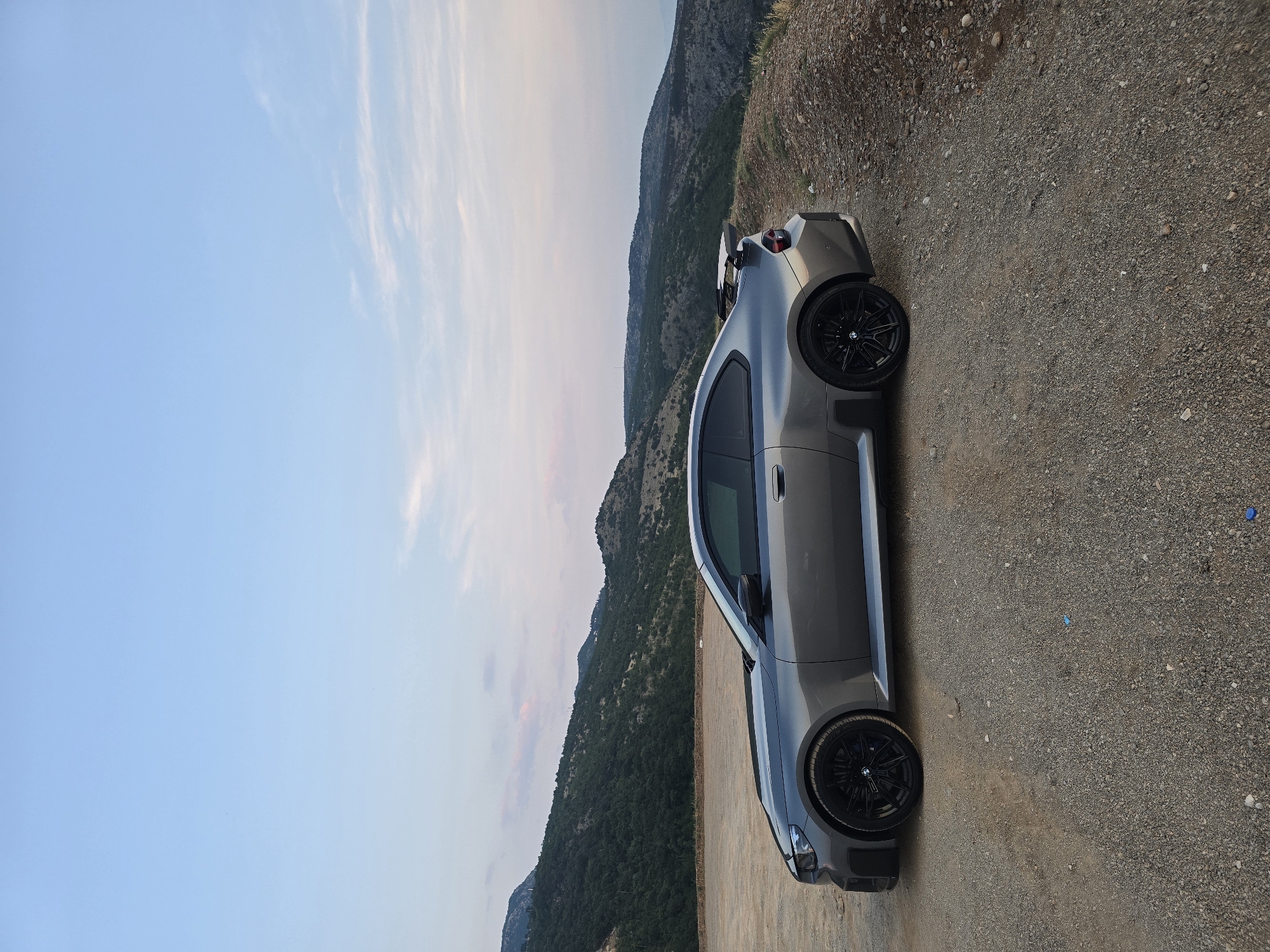 A gray BMW M2 Coupe is parked on a gravel surface overlooking a mountainous landscape. The car is positioned in the center of the frame, facing left, with its rear end towards the right. The car has black rims and a prominent rear spoiler. The background consists of rolling green hills and mountains under a pale blue sky with scattered clouds. The area appears to be a scenic overlook, possibly in a rural or mountainous region.