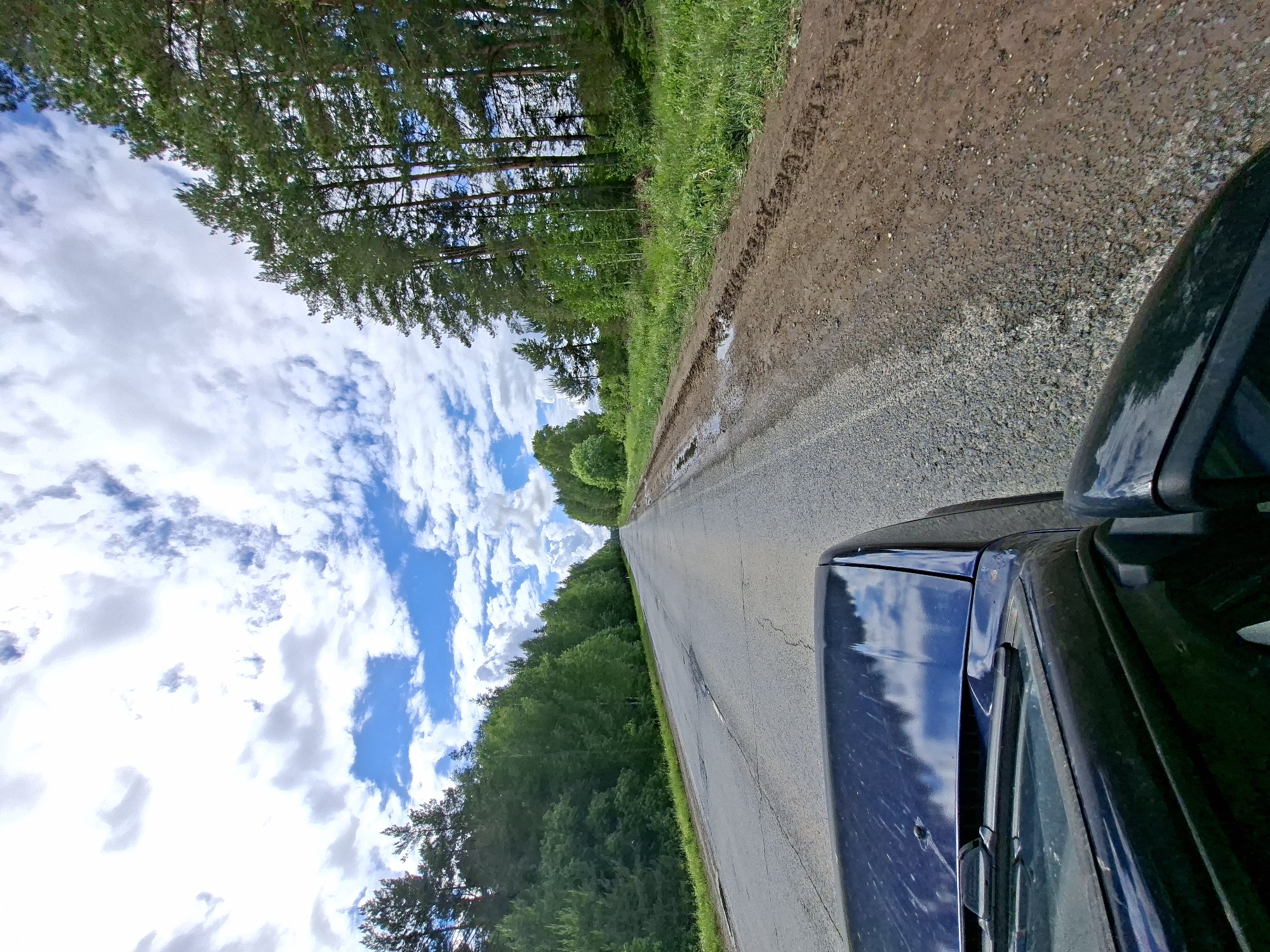 This image captures a view from a dark-colored vehicle on a rural road in Chumna, Russia, during daytime. The road, made of asphalt with some visible wear and cracks, stretches straight into the distance, flanked by dense forest.
In the foreground on the right, part of the vehicle, including its hood, windshield, and side mirror, is visible, reflecting the blue sky and trees. Adjacent to the road on the right is a dirt and gravel shoulder, appearing damp with mud and small puddles, suggesting recent rainfall. Beyond this shoulder, a thick line of green grass transitions into a dense forest composed of tall coniferous and deciduous trees, extending along both sides of the road and into the horizon.
Above the tree line, a bright blue sky is adorned with numerous fluffy white clouds, indicating clear weather. The scene conveys a sense of a journey through a peaceful, natural environment. No text is visible in the image.