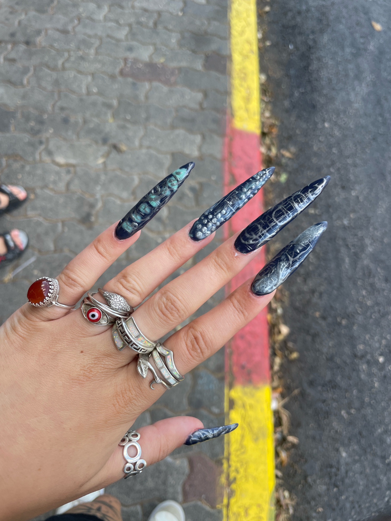 The image shows a close-up of a person's left hand, adorned with multiple rings and long, pointed fingernails painted with a dark, marbled design. The hand is positioned over a paved walkway next to a curb painted with red and yellow stripes, indicating an outdoor urban setting. The background shows asphalt and some dried leaves. The red and yellow striped curb suggests the location might be near a street or a designated parking area. The style of the paving stones and the curb paint are common in urban environments. While the image does not explicitly identify Raanana, Israel, the presence of similar street markings and paving styles can be found throughout the country.
