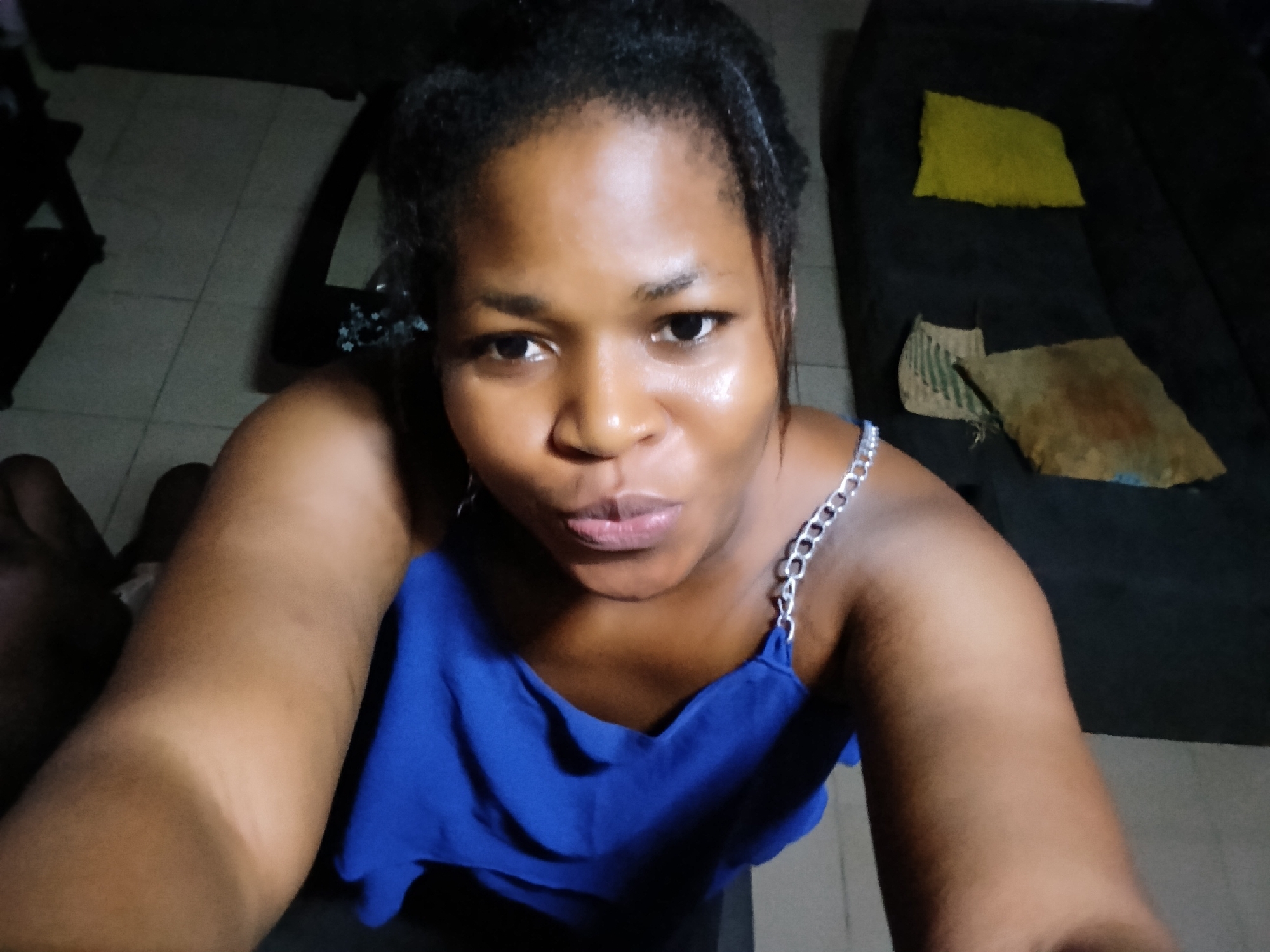 In an indoor setting, a Black female is taking a selfie. She is wearing a royal blue tank top with silver chain straps. Her hair is dark and pulled back. Her lips are pursed. Behind her, to the right, a dark sofa is visible with two throw pillows, one yellow and one brown. To the left, a tiled floor is visible. The lighting is dim.