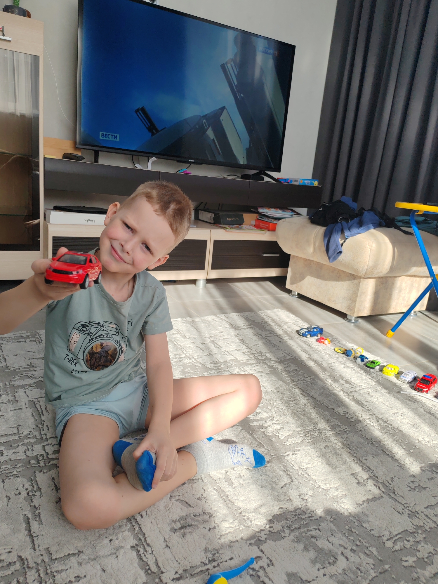 An indoor scene in Chaikovsky, Russia, depicts a young male child, approximately 6-8 years old, seated cross-legged on a light-colored, geometrically patterned rug. The child, with light brown hair, wears a light blue t-shirt featuring a camera graphic and light blue shorts, with grey socks. He holds a red toy car in his left hand, presenting it towards the viewer, and is smiling.
In the background, a large flat-screen television displays what appears to be a news program, evidenced by the "ВЕСТИ" and "РОССИЯ 1" logos, with imagery suggesting industrial or military content. Below the television is a dark media console. To the left, a light wood and glass cabinet is visible. To the right, dark grey curtains are drawn, and a beige upholstered footrest is present, topped with dark blue clothing. A blue and yellow toy stand or chair is also on the right.
A line of approximately 8-9 miniature toy cars, in various colors including blue, pink, yellow, green, white, and red, are arranged on the rug to the right of the child. Direct sunlight streams in from the right, casting distinct shadows and patterns across the rug. A blue and yellow toy piece rests on the rug in the lower foreground.