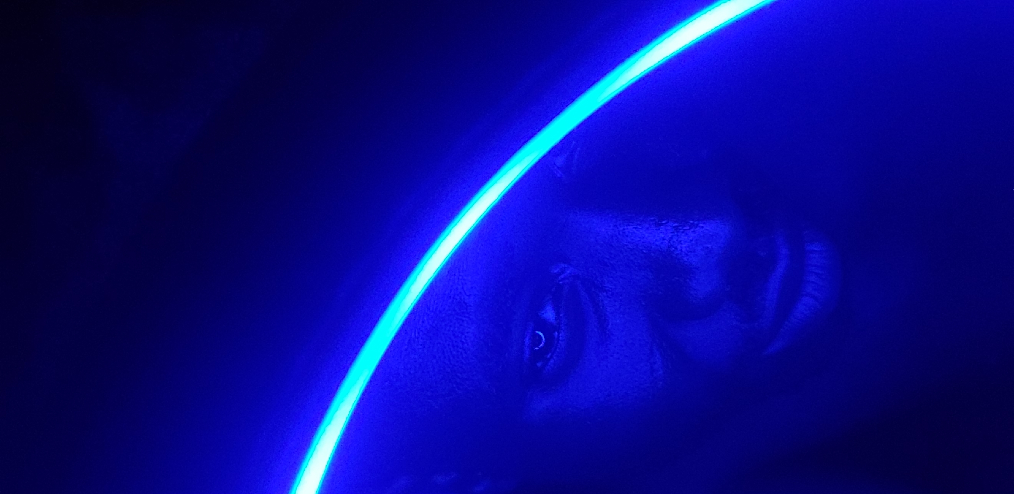 The image depicts a close-up of a person's face illuminated by a curved blue light source in a dark environment. The individual's features, including an eye and mouth, are partially visible. The scene is dominated by shades of blue. The location is not evident from the image and the mention of Narayi, Nigeria, is not supported by the visual content.