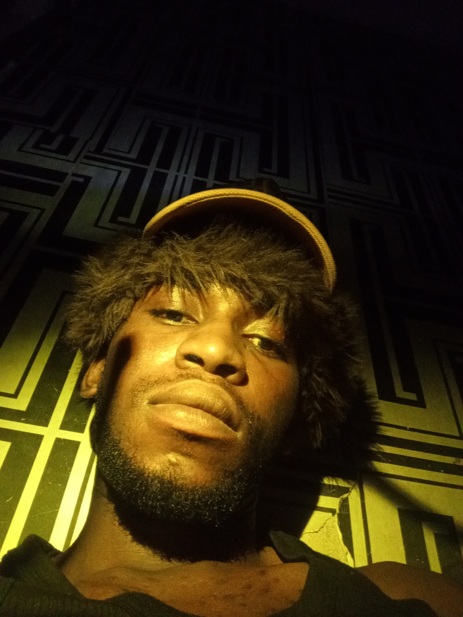 The image is a selfie of a young Black man wearing a furry hat and a dark t-shirt. He has dark skin, a short beard, and his eyes are looking directly at the camera. The background appears to be a wall with a geometric pattern, lit by a yellow light that casts shadows. The overall lighting is dim, creating a dramatic effect. The image appears to have been taken indoors. The location is not specified within the image or its metadata, and therefore cannot be determined to be in Lokoja, Nigeria.