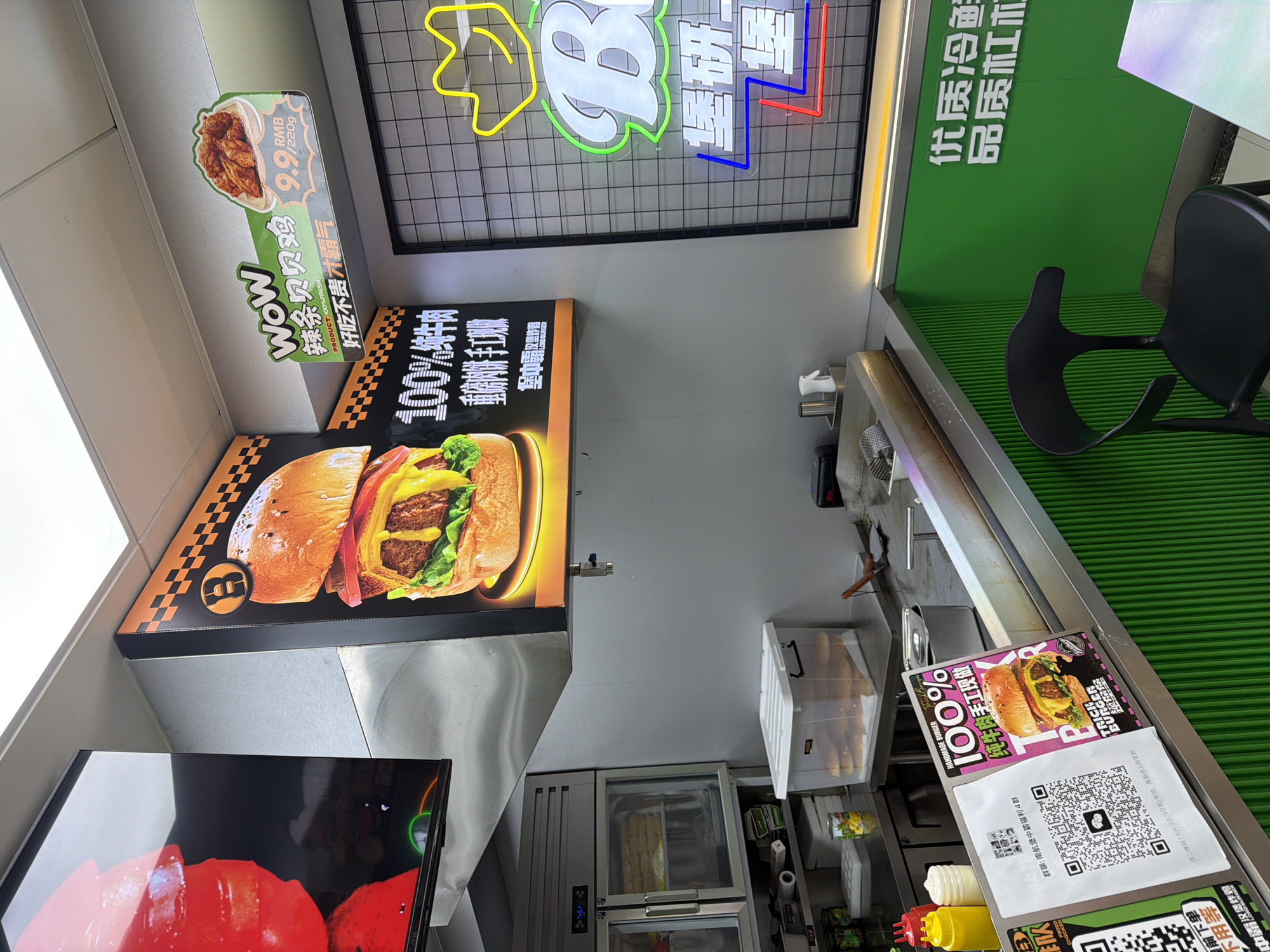 The image shows the interior of a "Trick Burger" restaurant in Jiangning District, China. A black chair sits next to a green wall with white Chinese characters. Above the food preparation area, signs advertise the restaurant's offerings, including "WoW" spicy chicken strips for 9.9 RMB and 100% beef burgers. A QR code is visible on a sign on the counter. The restaurant appears to be clean and well-lit, with ingredients stored in clear containers.