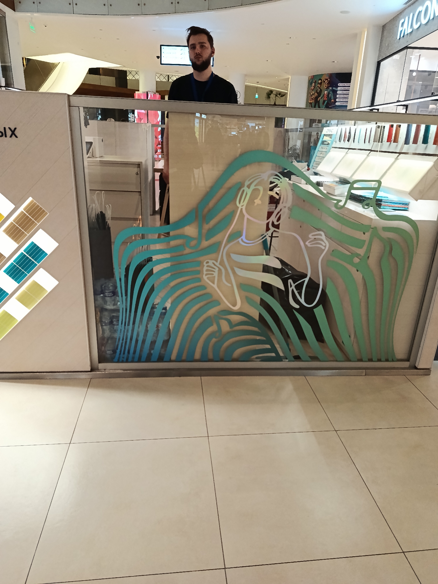 The image shows an indoor scene, possibly a shopping mall. There is a service counter or display case with a glass partition decorated with a stylized image of a woman wearing headphones. A man with a beard is standing behind the counter. The flooring is tiled and there are other shops and signage visible in the background. The lighting is bright and diffuse.