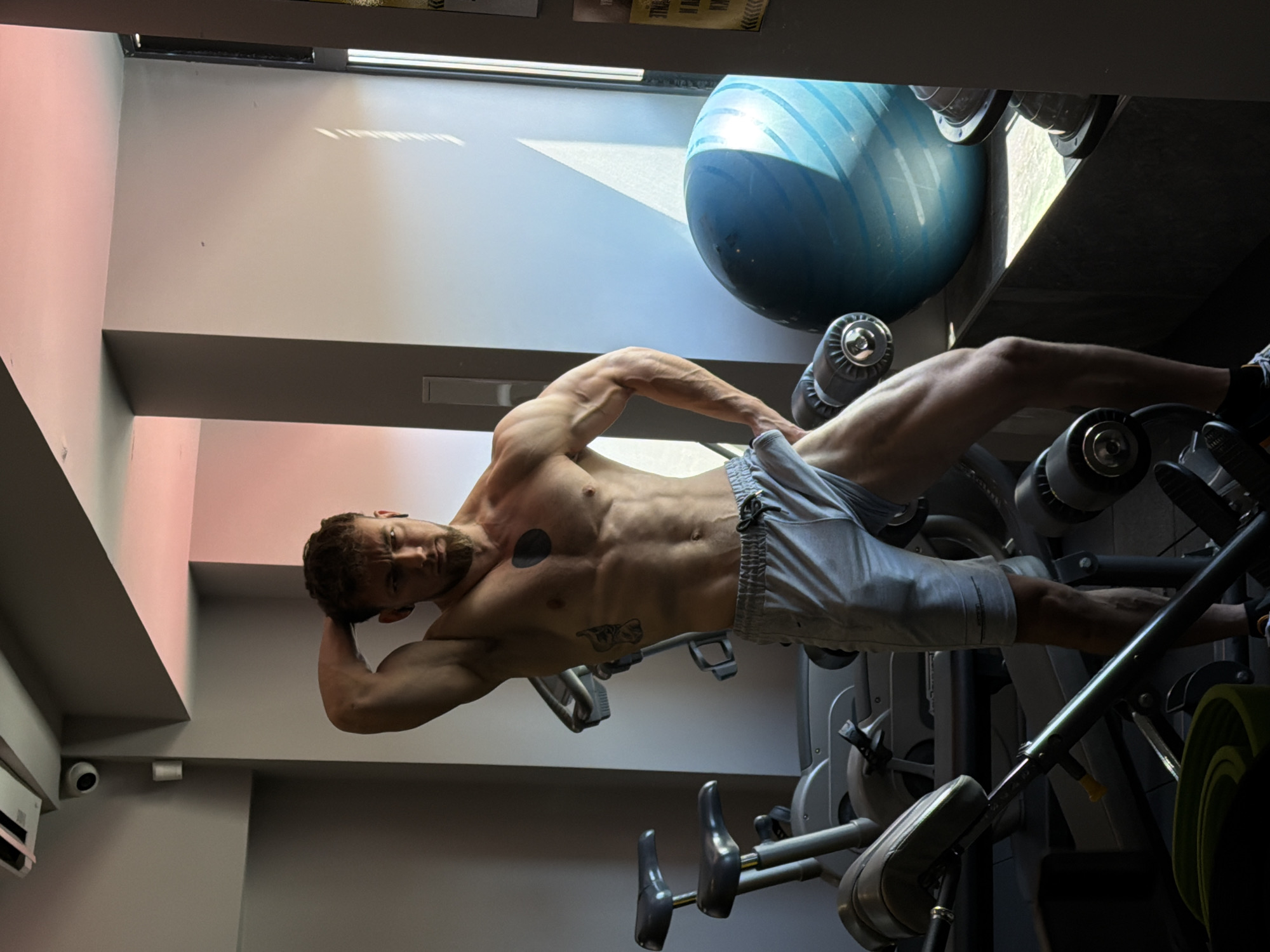 A muscular man in grey shorts stands in a gym in what appears to be Tsarevo, Bulgaria. He is shirtless, revealing tattoos on his torso. He is positioned near exercise equipment, including a weight machine and a blue exercise ball. Natural light streams through a window behind him. The gym has grey walls and a security camera is visible in the upper left corner.