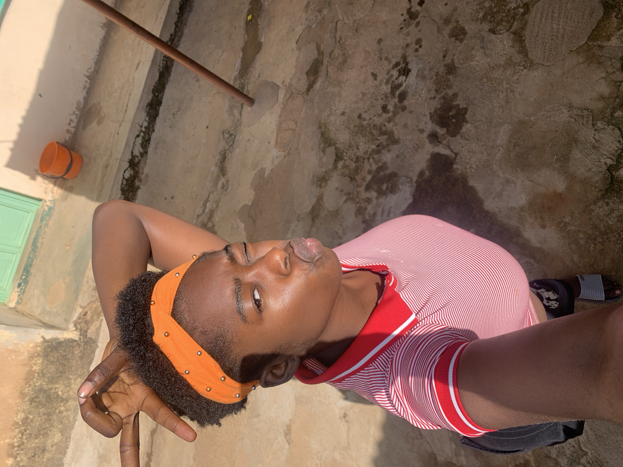 A young Black female is captured in a high-angle selfie outdoors, likely in Rafin Par, Nigeria. She is wearing a red and white striped polo shirt and an orange headband adorned with small gold beads. Her left hand is raised, forming a 'V' gesture near her head, and her right eye is winked. Her lips are pursed in a kissing expression. The ground beneath her appears to be a rough, weathered concrete surface, with some darker stains and cracks. In the background, a light-colored wall with a green-framed window and an orange bucket are visible. A wooden pole stands upright on the right side of the frame. The lighting suggests it is a sunny day, casting shadows.