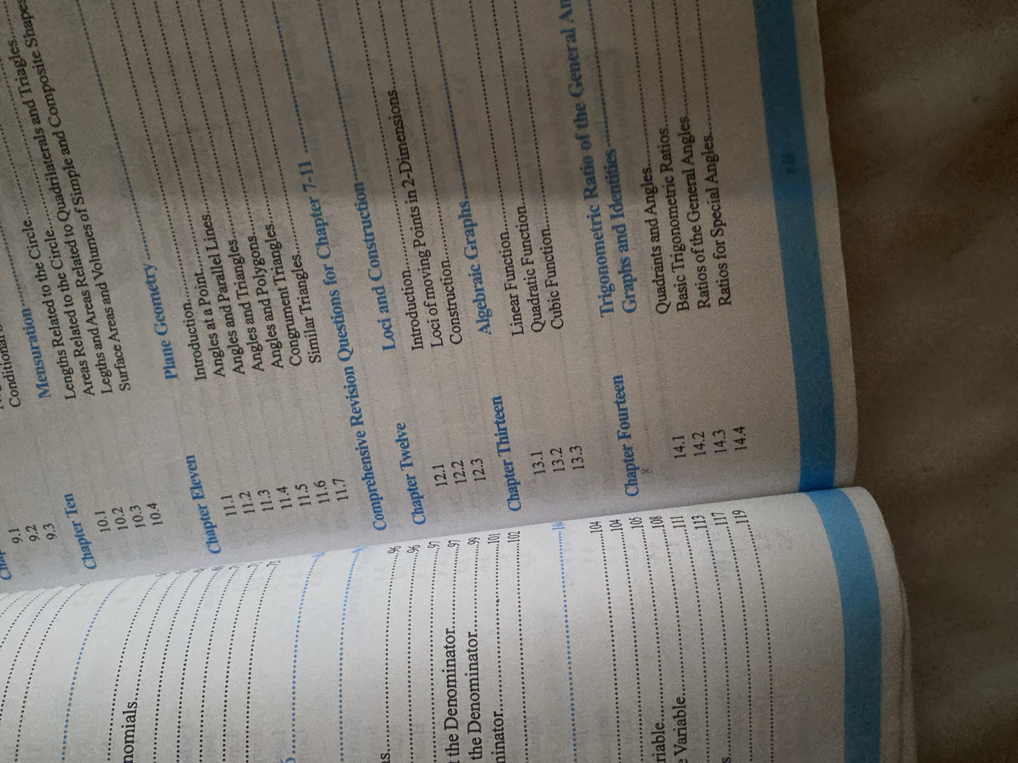 The image is a close-up photograph of an open book, displaying a table of contents. The left page features a list of topics related to "nominals", "the Denominator", and "riable". The right page lists chapters such as "Mensuration", "Plane Geometry", "Comprehensive Revision Questions for Chapter 7-11", "Loci and Construction", "Algebraic Graphs", "Trigonometric Ratio of the General Angles and Identities", "Chapter Ten", "Chapter Eleven", "Chapter Twelve", "Chapter Thirteen", and "Chapter Fourteen". The book is lying on a textured surface, possibly a table or floor, with a bright blue and white binding visible at the bottom. The lighting suggests an indoor setting, and the image quality is clear, allowing for easy readability of the text. There are no people, city elements, or other environmental features visible in the image. The location of Jalingo, Nigeria is not discernible from the visual information present in the photograph.