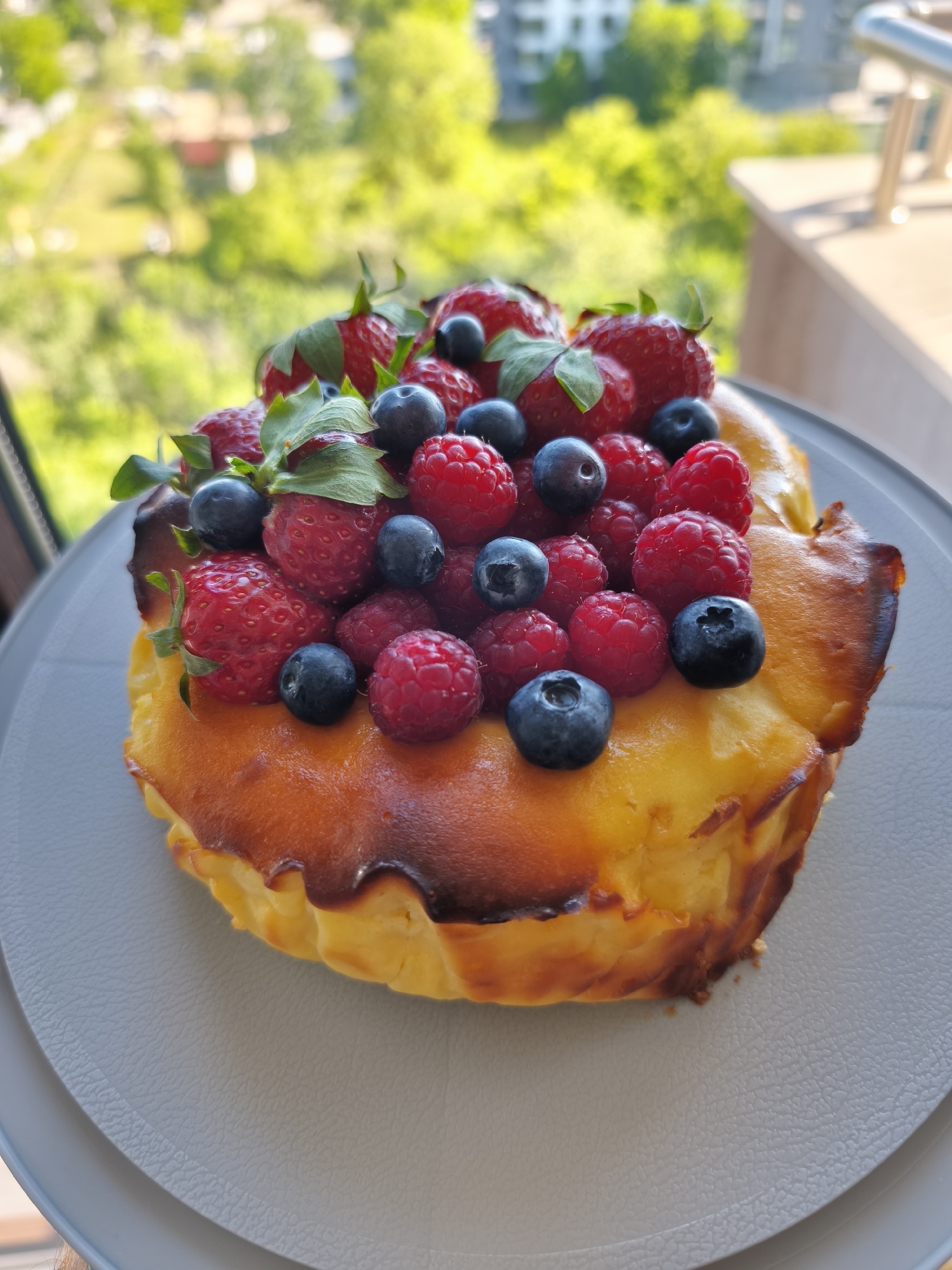 A close-up shot shows a baked dessert, likely a cheesecake, garnished with fresh strawberries, raspberries, and blueberries. The dessert is slightly burnt on the edges and is placed on a light gray plate. The background is blurred, showing trees, buildings, and what appears to be a balcony railing, suggesting the photo was taken indoors with a view of an urban area. Based on the prompt, the location is Plovdiv, Bulgaria. No people are visible in the image. The interaction is primarily between the dessert and its setting, where the dessert is the main subject against the backdrop of the city.