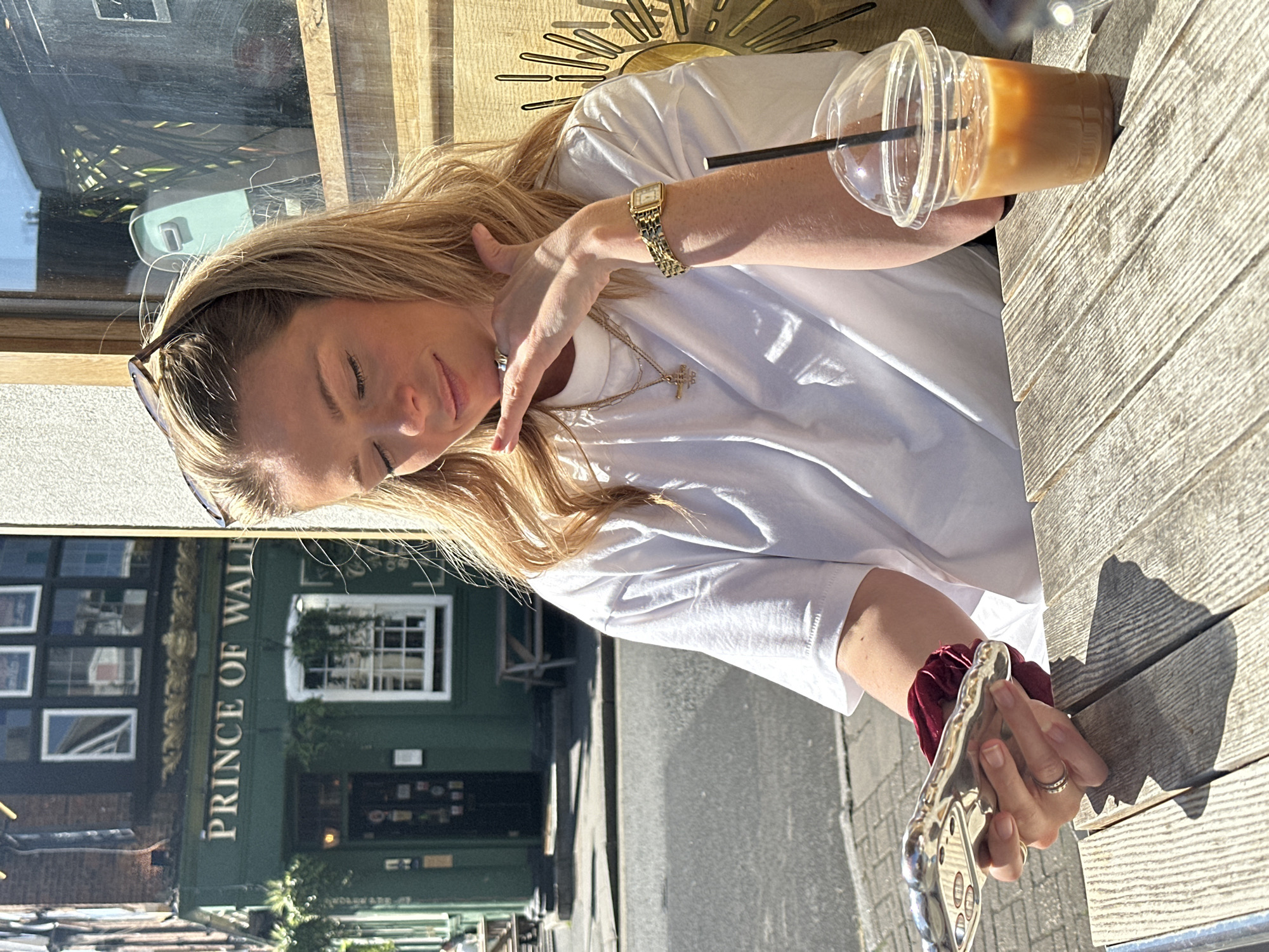 A blonde woman wearing a white t-shirt is seated at an outdoor wooden table at a cafe. She has sunglasses on top of her head, and a gold watch and necklace are visible. She is holding a mobile phone in her right hand and touching her neck with her left hand. An iced coffee drink sits on the table. The "Prince of Wales" pub is visible in the background, indicating the location is likely in London, United Kingdom. The weather appears sunny.