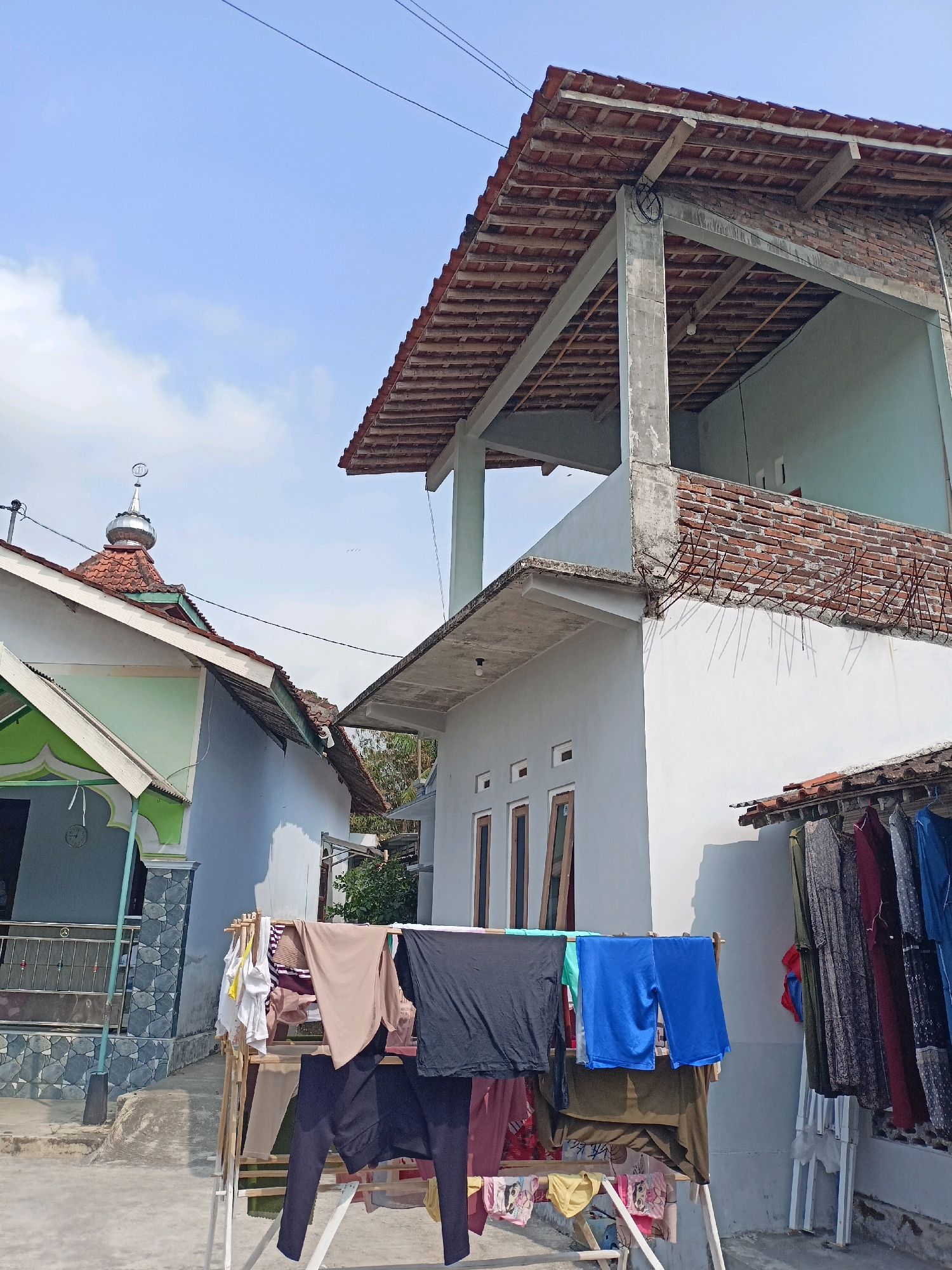In Bandongan, Indonesia, an outdoor scene captures laundry drying under a clear sky. A clothesline in the foreground holds a variety of garments, including a black t-shirt, blue shorts, and several other items of clothing. To the left, a building with a light blue exterior and a green awning is partially visible, featuring a dome-shaped roof and a crescent moon ornament. Adjacent to it, a multi-story building with unfinished brickwork on the upper level and a white facade on the lower level is under construction. The lower part of this building has several windows. To the right, more clothing is hanging on a clothesline attached to a structure, with several dresses visible. The ground appears to be paved. Overhead, power lines crisscross the sky, and a few birds are in flight.