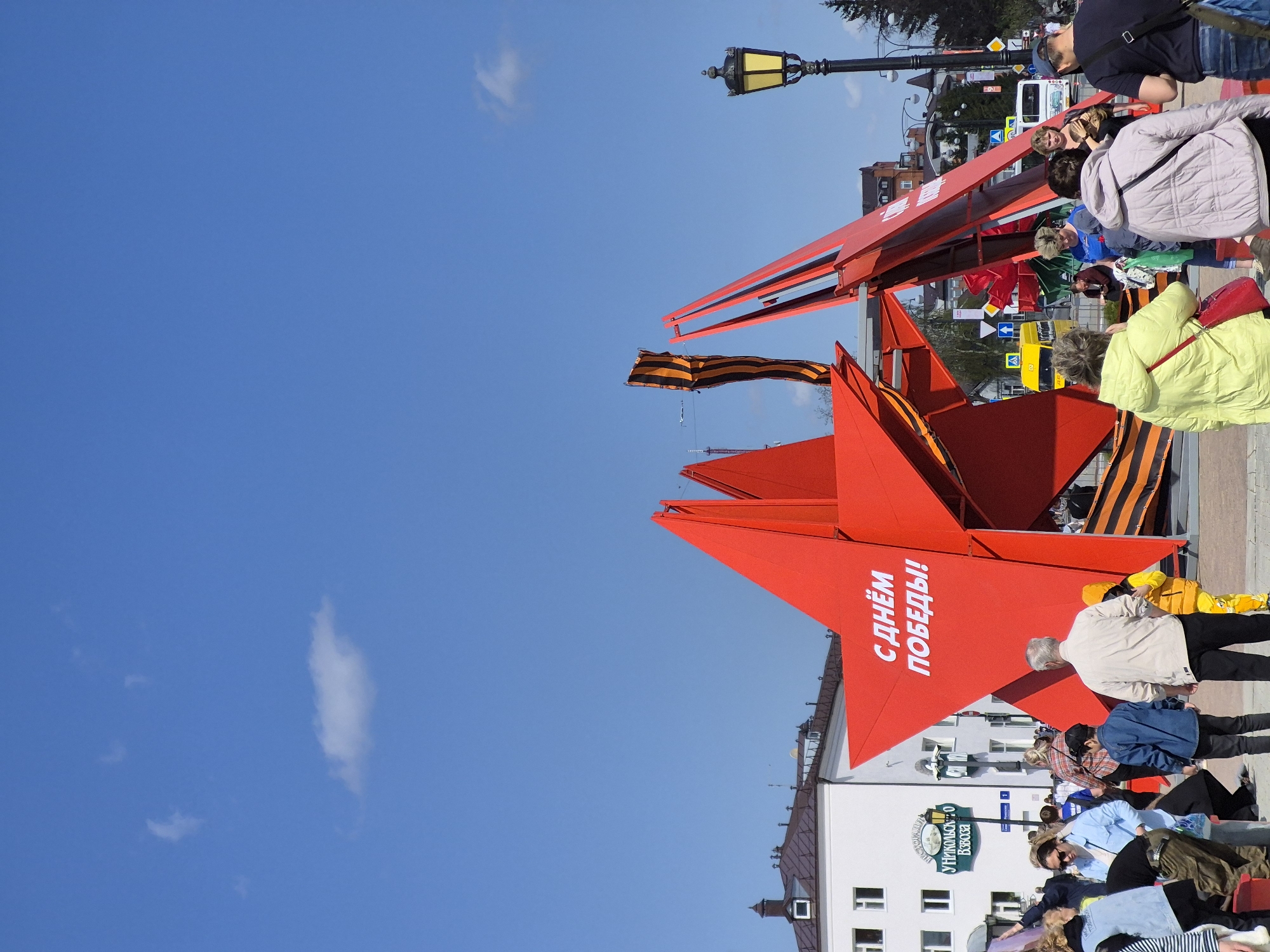 The image shows a public celebration in Tobolsk, Russia, featuring red star decorations emblazoned with "С Днём Победы!" (Happy Victory Day!) and St. George's ribbon, commemorating Victory Day. Several people are visible, walking around or sitting. The sky is clear blue with a few scattered clouds. The architecture is visible, including buildings and streetlamps.