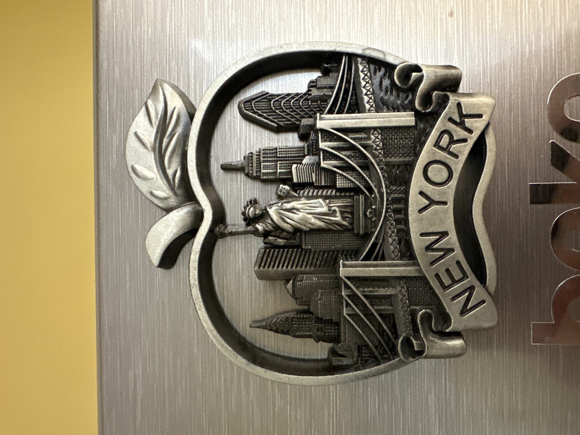 A close-up, slightly rotated shot captures a metallic relief art piece, likely a souvenir magnet, celebrating New York City. The central object is shaped like an apple, a nod to "The Big Apple" nickname, complete with a detailed leaf stem on its left side.
Within the apple's outline, various iconic New York City landmarks are intricately depicted in a three-dimensional, antiqued silver or pewter-colored metal. Prominently featured is the Statue of Liberty, situated in the lower-middle section, facing towards the right. Surrounding the statue and filling the upper and lower sections are several recognizable skyscrapers, including what appears to be the pointed spire of the Chrysler Building and other city structures. A large bridge, possibly the Brooklyn or Manhattan Bridge, stretches across the upper right portion of the apple.
A banner-like scroll runs vertically along the right side of the apple, bearing the embossed text "NEW YORK" in capital letters. The entire relief has a textured, slightly worn appearance, enhancing its classic appeal.
The metallic art piece is mounted against a background of brushed silver-colored metal, characterized by distinct vertical striations. To the far left, a solid, plain yellow surface is visible, contrasting with the metallic textures. To the right of the apple, partially cropped, are fragments of large, metallic letters, possibly "YO" from another word or display. The lighting is soft and even, highlighting the details of the relief without harsh shadows. No specific activity or event is taking place; the image focuses solely on the decorative object. The precise location is unknown, but the background suggests an indoor setting.