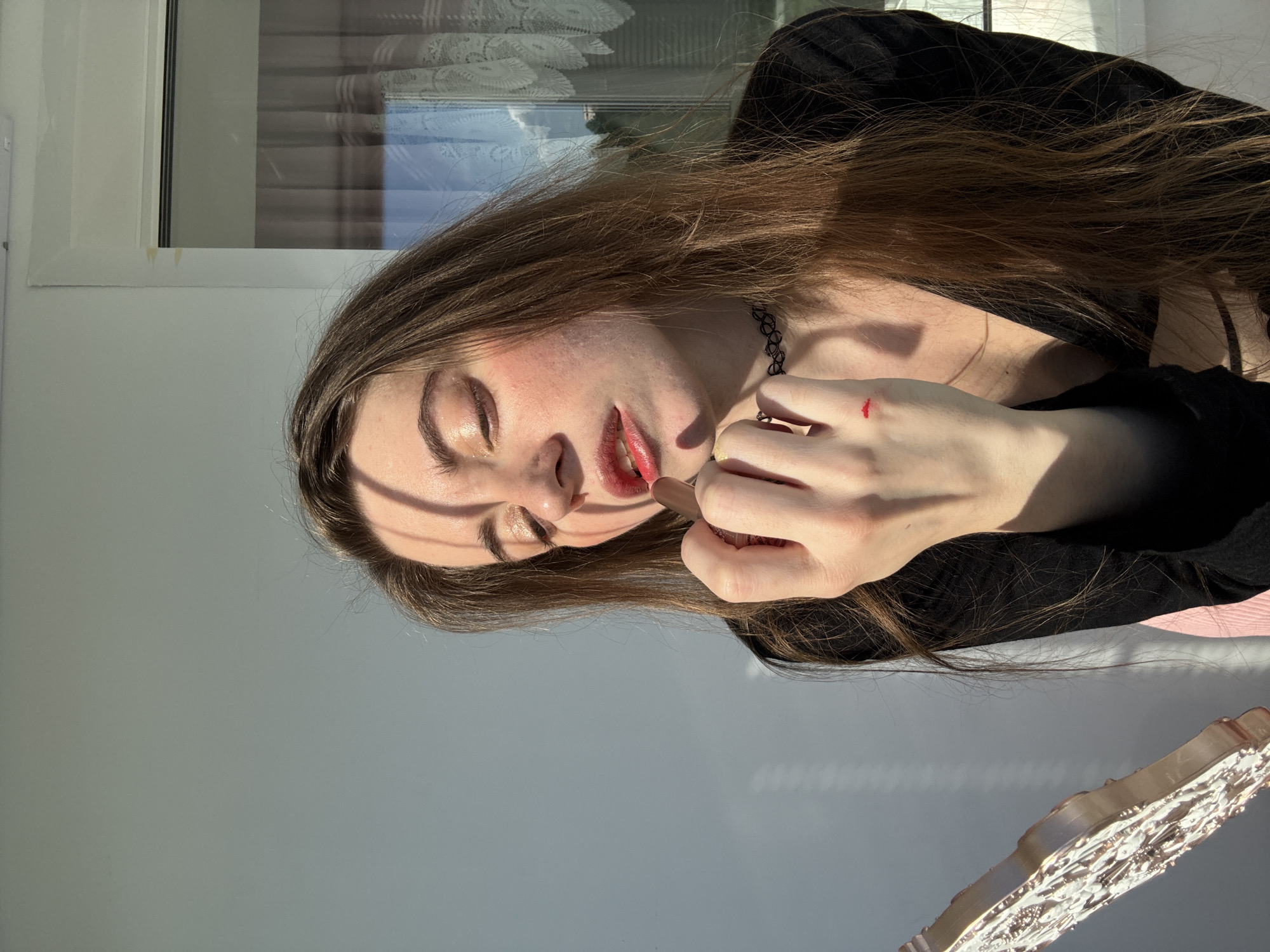 The image shows a young woman applying lipstick. She has long, light brown hair and is wearing a black top. Her eyes are closed, and her mouth is slightly open as she applies a pale pink lipstick. There is a thin choker necklace around her neck. Her right hand is raised to her face, and there is a small red mark on the back of her hand, possibly from makeup. The lighting suggests it is daytime, with strong sunlight casting shadows across her face and the background. The background consists of a pale grey wall and a window with white blinds. A decorative silver object is visible in the lower right corner of the image. The overall mood appears to be one of concentration or relaxation.