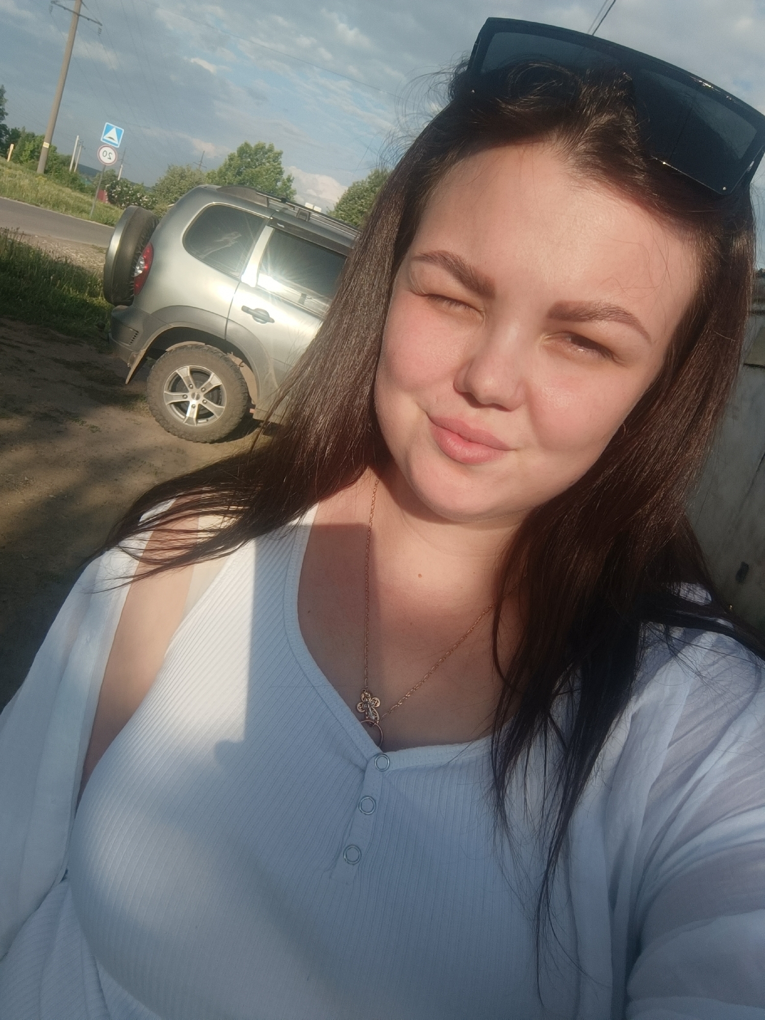 A selfie shows a woman with dark hair, wearing a white top and a gold necklace with a cross pendant. She is standing outside, with sunlight shining on her face, causing her to squint one eye. Sunglasses are perched on top of her head.
In the background, a silver SUV with a spare tire mounted on the rear is parked near a road, where a road sign is visible. The setting appears to be an urban area, possibly a residential neighborhood or a suburban area with trees and a utility pole visible in the distance.
Based on the metadata, the image was taken in Zav'yalovo, Russia.