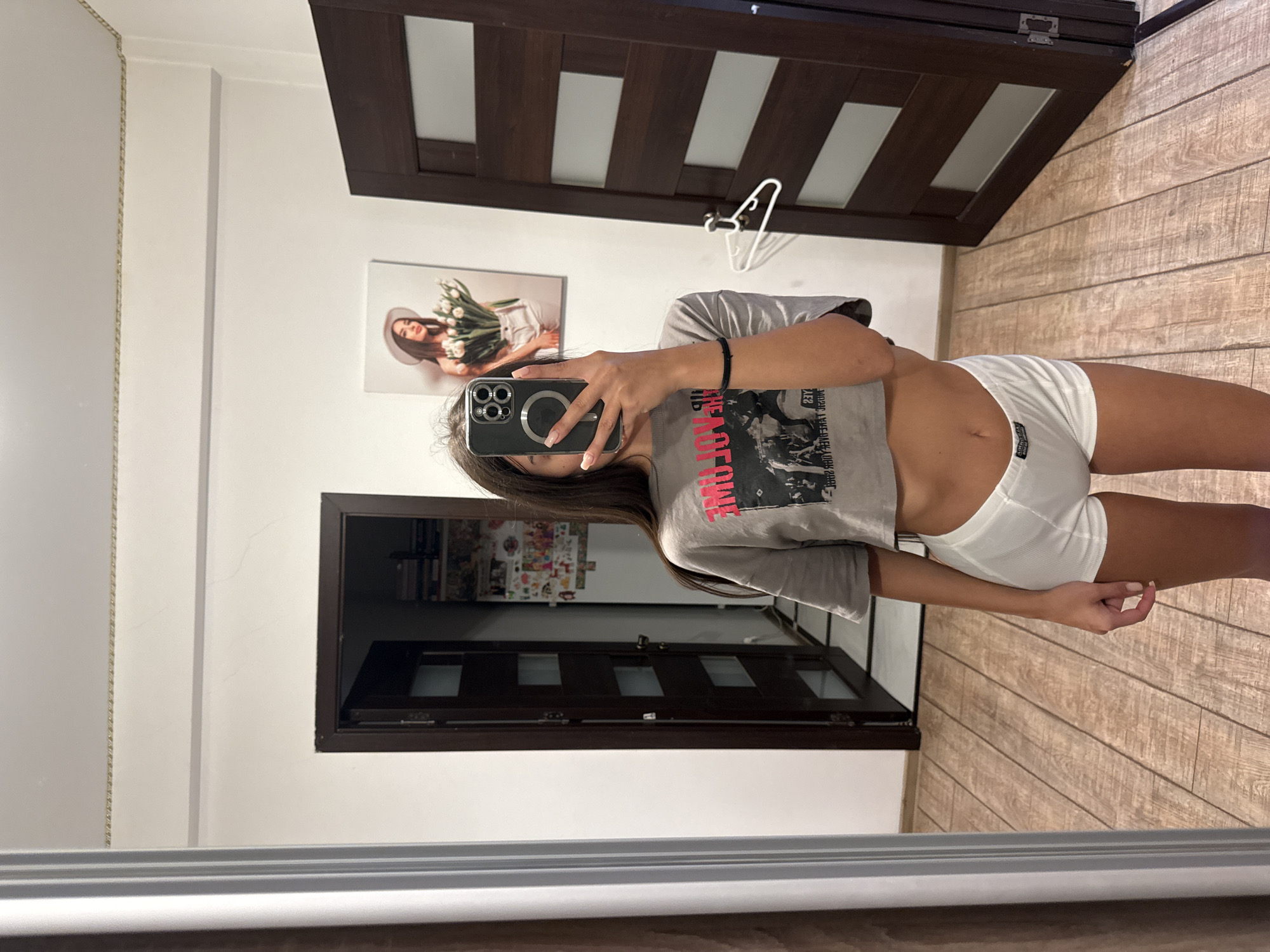 The image shows a young woman taking a mirror selfie in a room. She is wearing a grey crop top with a graphic print and white high-waisted shorts. The room appears to be a hallway or entryway with wooden floors and dark wood doors. There is a framed picture of a woman with flowers on the wall. A mirror is positioned to reflect the woman and part of the room. The overall impression is a casual, indoor setting.