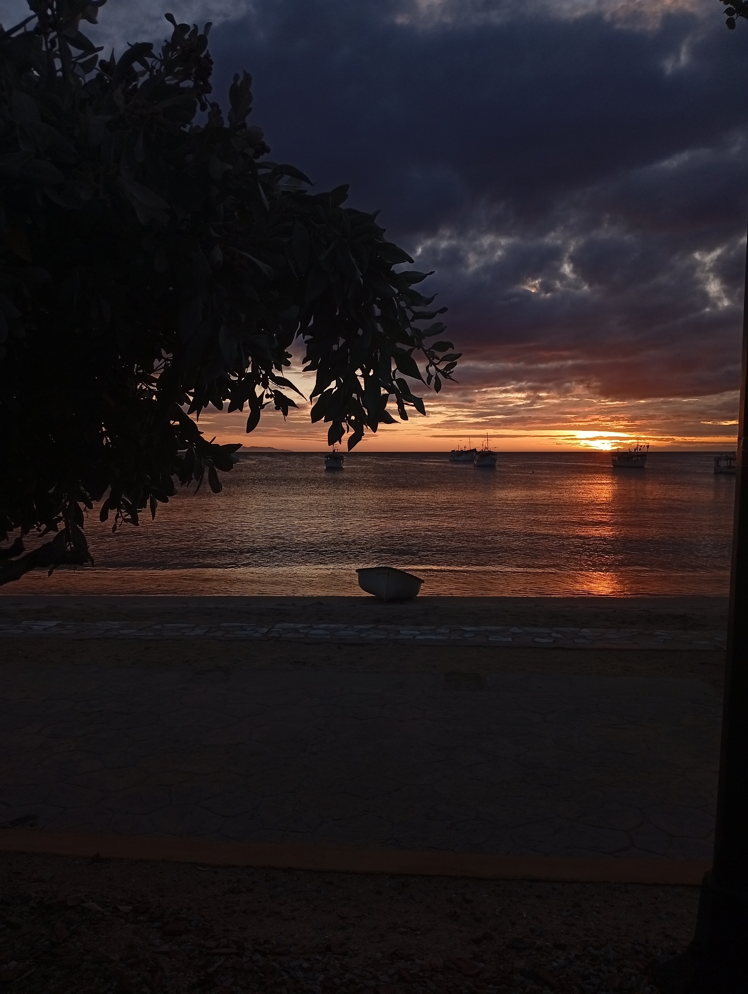 The image depicts a serene yet dramatic coastal sunset scene. The sun, a bright orange orb, is setting on the horizon, casting brilliant warm hues of orange, red, and gold across the lower sky. These vibrant colors are intensely reflected on the surface of the calm water, creating a shimmering path of light that extends towards the viewer.
Above the horizon, dark, heavy clouds dominate the sky, ranging in color from deep purples and grays to dark blues. These clouds create a stark contrast with the illuminated horizon, suggesting an overcast evening or perhaps the aftermath of a storm. Sunlight occasionally breaks through, highlighting the edges of some clouds.
Several boats are visible on the water, silhouetted against the bright sunset. These appear to be medium-sized vessels, possibly fishing boats, anchored or drifting peacefully. In the foreground on the left, a large, dense tree or bush is heavily silhouetted, its dark foliage framing the scene. The immediate foreground features a dark shoreline or beach, with a section of a path or low wall made of irregular stone or concrete slabs. A small, light-colored boat is resting on the sand near the water's edge, appearing to be overturned or lying on its side. In the far distance, a faint outline of land or an island is visible on the left horizon.
The overall mood is peaceful and contemplative, with the dramatic sky adding a touch of grandeur to the tranquil setting. No people or visible text are present in the image.