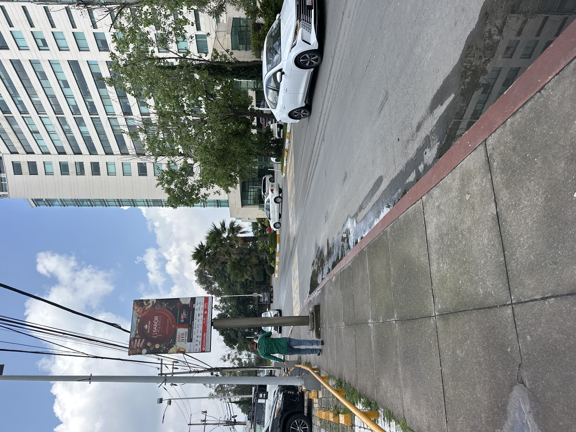 A lone man wearing a green shirt and blue jeans walks on a concrete sidewalk next to a street in the city of Jesús del Monte, Mexico. The man is facing away from the camera and is carrying a brown backpack on his left shoulder. To his left, a billboard is mounted on a pole. The billboard advertises a "Sabor Polanco" event featuring food and beverages, with dates in March. A white sedan is driving on the street, passing by a large, modern white building with many windows. Trees and palm trees are visible in front of the building. The street has puddles of water on it, reflecting the sky and the building. The sidewalk is lined with yellow bollards and a red curb. The sky is cloudy with patches of blue.
