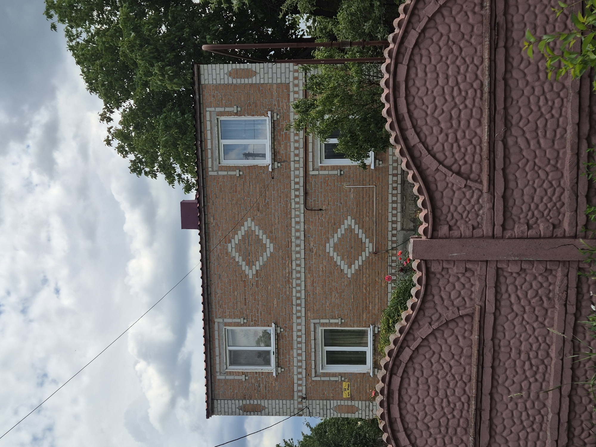 This is a vertical, eye-level, medium shot of a two-story brick house and a portion of a decorative gate. The house is constructed of reddish-brown bricks with lighter-colored bricks used for decorative accents. These accents include vertical strips flanking the windows and diamond shapes patterned on the upper section of the facade. There are four windows visible, two on each floor, all with white frames. The upper windows reflect a cloudy sky, while the lower windows show green curtains. A dark red metal roof is visible along the top of the house. To the right, a large, ornate gate made of reddish-brown concrete takes up a significant portion of the frame. The gate's surface is textured with a pattern resembling rounded stones, and it features a curved decorative element on the left side. Some green foliage and a few red flowers are visible behind the gate and to the left of the house. A thin electrical wire stretches diagonally across the sky. The sky is filled with white and gray clouds, suggesting overcast weather, possibly in the late afternoon or early evening. A yellow street sign with dark text is affixed to the house wall near the lower right window. The text on the sign appears to be "вул. Миру" which translates to "Myru Street". The scene is likely a residential area in Sozonivka, Ukraine. No people are visible in the image.