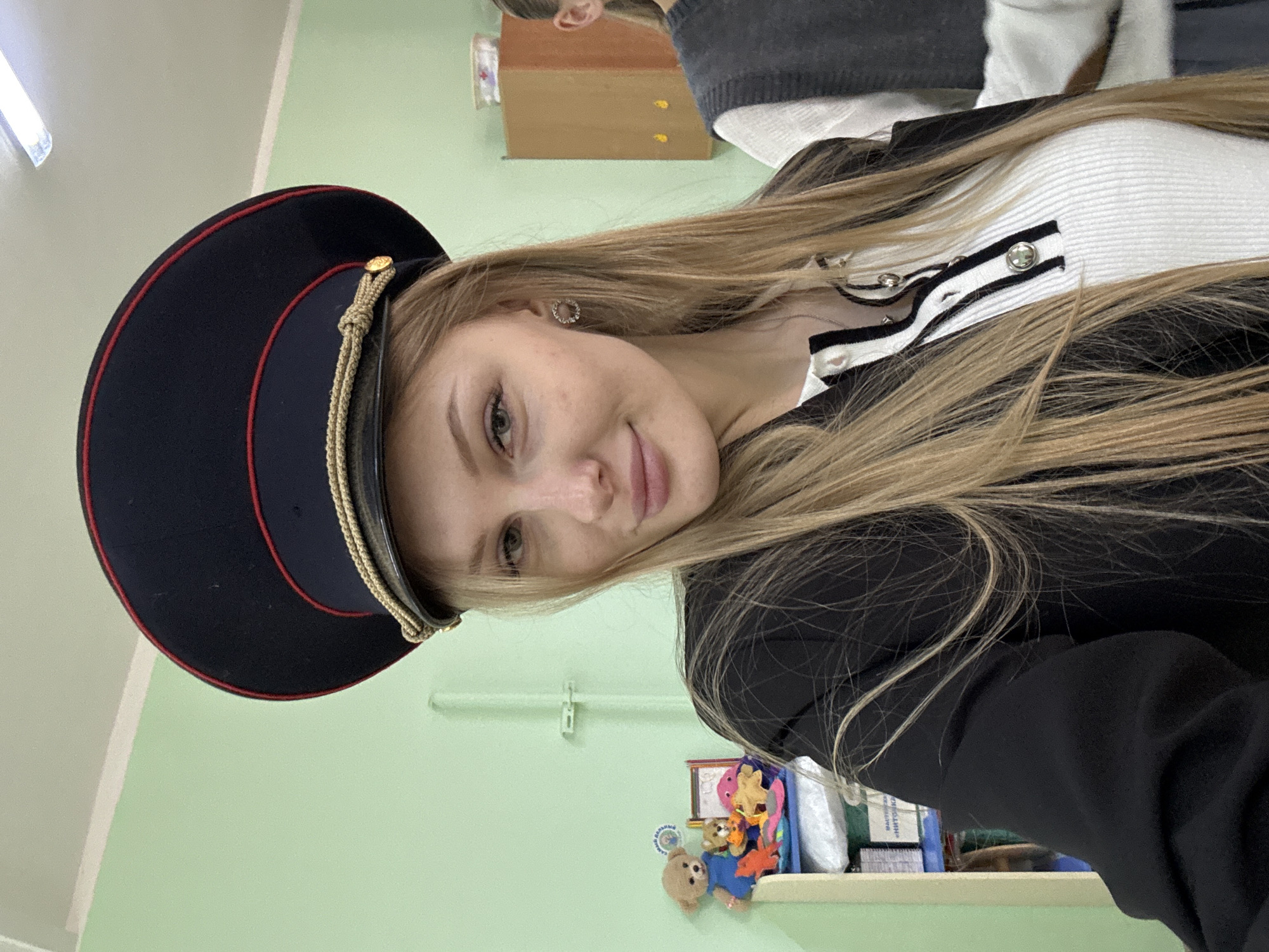 A young woman is posing indoors. She is wearing a black peaked cap with a red trim and gold cord, a black blazer, and a white top with black and white stripes. She has long blonde hair and is wearing earrings. The background is a light green wall with a light switch and a shelf with various small toys. A piece of wooden furniture is partially visible in the background. The location may be in the city of Perm, Russia.