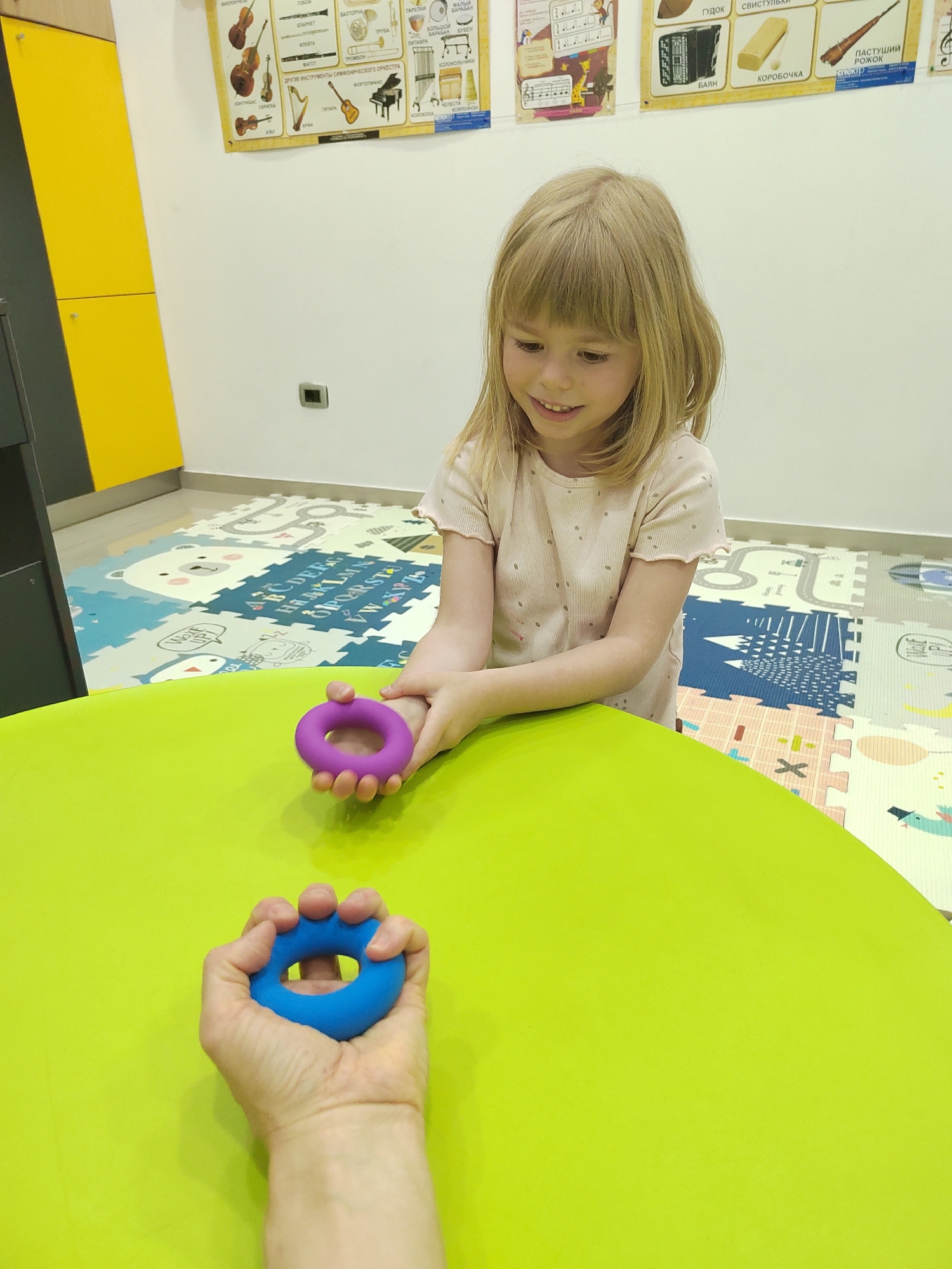 A young blonde-haired girl, with a gentle smile, is seated at a bright green table, holding a purple ring-shaped object with both hands. Opposite her, an adult's hand is visible in the foreground, holding a similar blue ring-shaped object. This suggests an interactive activity or play session, possibly involving hand exercisers or sensory toys. The girl is wearing a light-colored, short-sleeved top.
The setting is an indoor, well-lit child-friendly environment, likely a play area or educational facility in Podgorica, Montenegro. The floor is covered with a colorful foam playmat depicting various shapes, cartoon animals (including a bear and a whale), and sections of the English alphabet.
The white wall in the background features several educational posters. These posters illustrate various musical instruments and display text primarily in Cyrillic, identifying instruments such as "БАЯН" (bayan/accordion), "КОРОБОЧКА" (wood block), "ПАСТУШИЙ РОЖОК" (shepherd's horn), "ГУДОК" (whistle), "ТАПЕЛКИ" (cymbals), "БАРАБАН" (drum), "БОЛЬШОЙ БАРАБАН" (bass drum), "БУБЕН" (tambourine), "КОЛОКОЛЬЧИКИ" (bells), "КОНТРАБАС" (contrabass), "КЛАРНЕТ" (clarinet), "ФЛЕЙТА" (flute), "ТРОМБОН" (trombone), "ФОРТЕПИАНО" (piano), and "АРФА" (harp). One poster is titled "ДРУГИЕ ИНСТРУМЕНТЫ СИМФОНИЧЕСКОГО ОРКЕСТРА" (Other instruments of the symphony orchestra). A black electrical outlet is visible on the wall. The atmosphere appears calm and engaging, with the girl showing clear enjoyment.