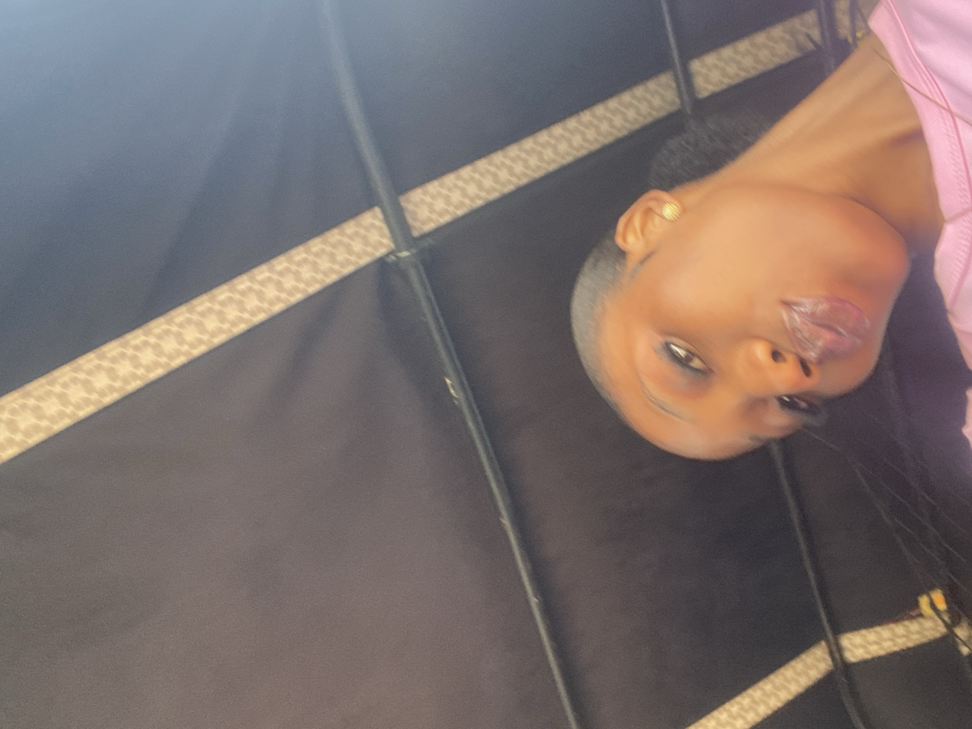 The image shows a low-angle, close-up selfie of a young Black woman with her head tilted and lips pursed. She is wearing a pink shirt and a gold earring. Her hair is dark and pulled back. The background appears to be the interior of a vehicle, possibly a bus or a rickshaw, with a dark fabric ceiling and a light-colored patterned trim running along the top. The lighting suggests it is daytime. The image quality is somewhat grainy, and the focus is on the woman's face. There are no specific landmarks or environmental cues that would definitively place the scene in Bar Jirgi Summa, Nigeria, or any other specific location.