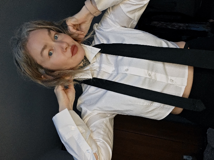 The image captures a waist-up shot of a person, likely a woman, looking directly at the camera with a slightly tilted head. The image itself is rotated, making the subject appear to be leaning to the right.
The person has light-colored hair, possibly blonde or light brown with gray strands, styled loosely around their face. Their eyes are open wide and appear blue. They have defined eyebrows and are wearing red lipstick. A dark, possibly tattooed, pattern is visible on their neck, extending from beneath their chin down towards the collarbone.
They are wearing a crisp white button-down shirt with the top few buttons undone, revealing a hint of skin on the chest area. The shirt has long sleeves with buttoned cuffs, and the cuffs are visible around their wrists. Over the white shirt, a black tie is worn loosely, with its two ends hanging down and partially obscuring the lower part of the shirt. The tie appears to be a solid black color. The shirt seems to be cropped, revealing a small sliver of midriff just above a dark piece of clothing, possibly trousers or a skirt.
The person's hands are raised, with their fingers lightly touching the hair near their temples or ears, as if adjusting their hair or posing. A light-colored watch or bracelet is visible on their left wrist.
The background is simple and appears to be an indoor setting. To the left, there's a dark gray or charcoal-colored wall. To the right and beneath the person, there's a dark, possibly wooden, surface that could be furniture like a dresser or a bed frame. The lighting suggests an indoor environment, possibly with artificial light. There is no discernible text visible in the image. The overall mood appears confident and direct, with a hint of playful posing.