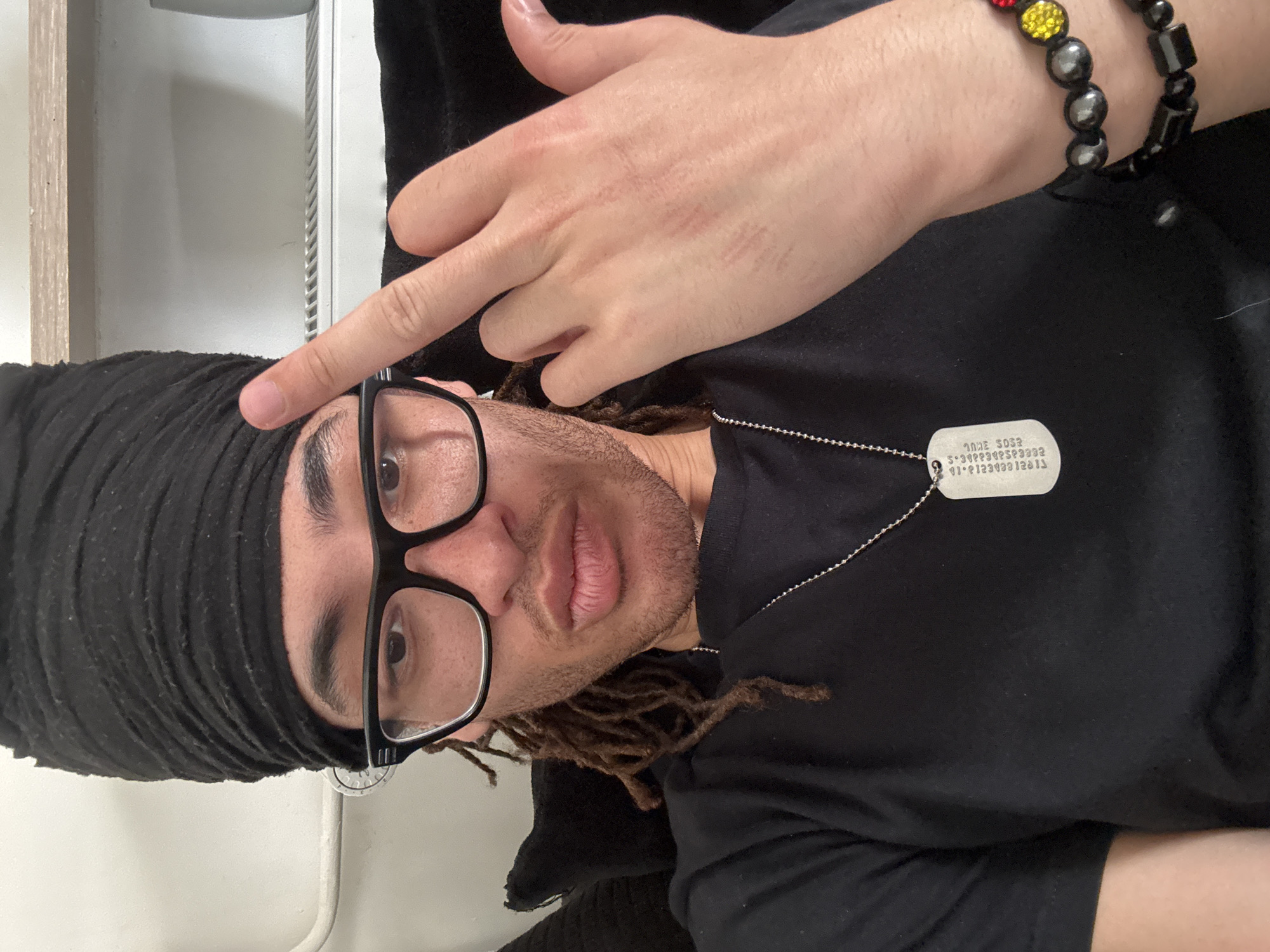 A person wearing a black headscarf, black-rimmed glasses, and a black t-shirt is visible. They have brown dreadlocks. A hand with a bracelet made of black, red, and yellow beads is raised, with the index finger pointing towards the person's forehead. A dog tag hangs from a ball chain around the person's neck. The background appears to be an interior space with white walls and a beige trim. There is no indication of the location being in the city of Paris, France.