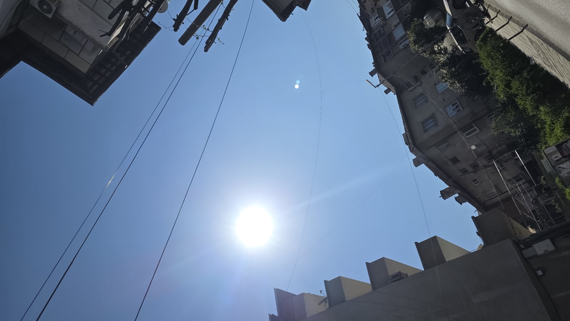 The image depicts a low-angle view of the sky in what is likely an urban environment. The bright sun dominates the lower center of the frame. Several buildings are visible around the edges of the frame. Electrical or communication wires crisscross the sky between the buildings. The image was taken in Blagoevgrad, Bulgaria.