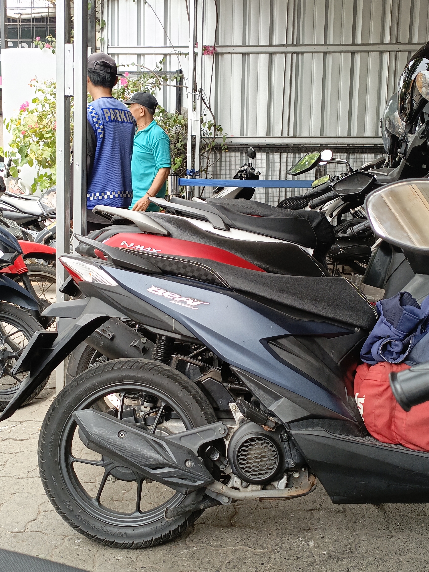 The image depicts a close-up view of several parked motorcycles in what appears to be a parking area in East Jakarta, Indonesia. In the background, two men are visible, one wearing a blue vest with the word "PARKING" on it and a dark cap, and the other in a turquoise shirt and a black cap. They are standing in front of a corrugated metal wall, with some greenery and flowers to their left. The motorcycles are the primary focus, with their seats, wheels, and parts of their engines clearly visible. One motorcycle in the foreground has a dark blue body and a prominent exhaust pipe. Red and black bags are piled on the seat of one of the motorcycles. The ground is paved with concrete.